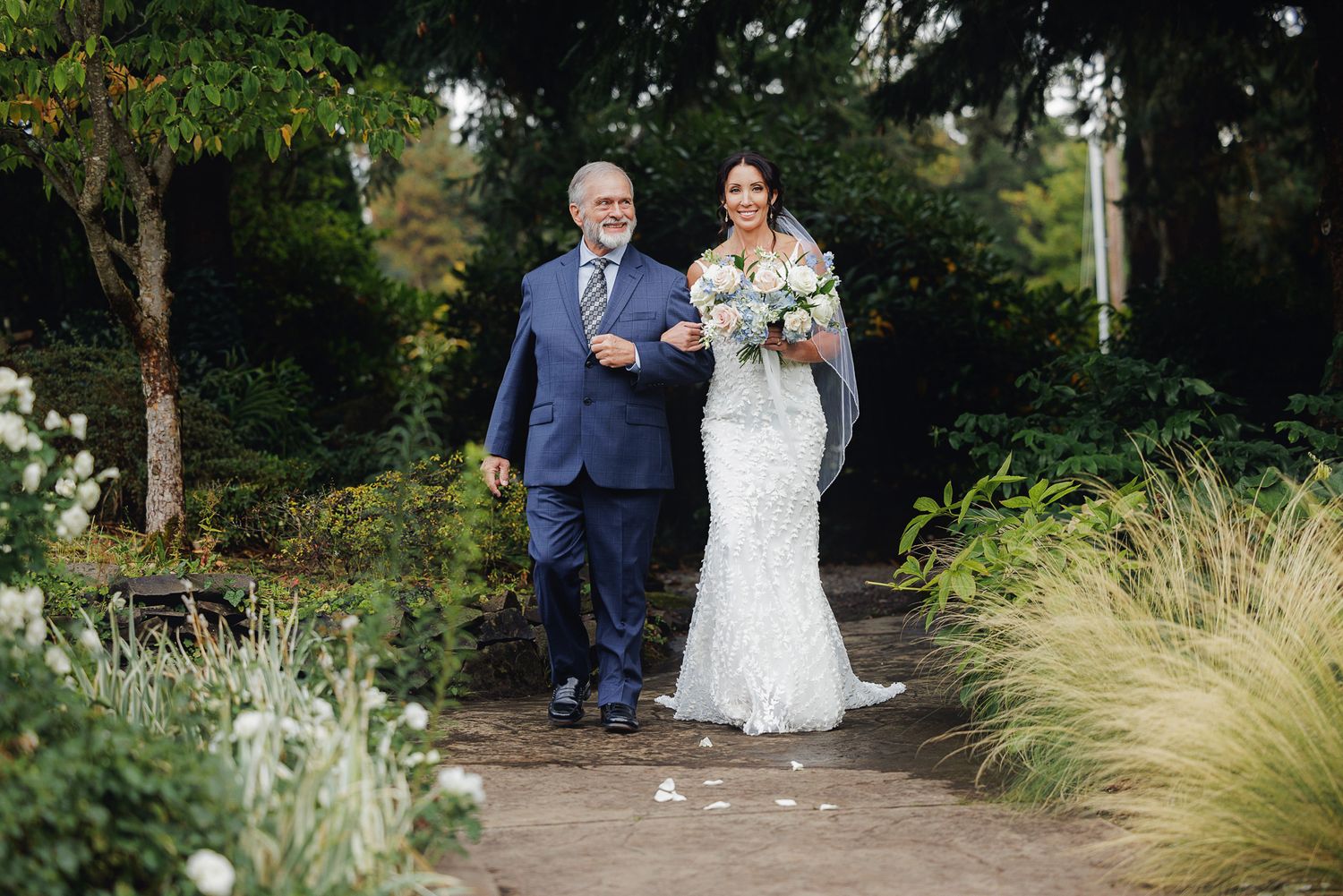 A bride in a lace mermaid gown walks down a garden path with an escort in a navy suit.