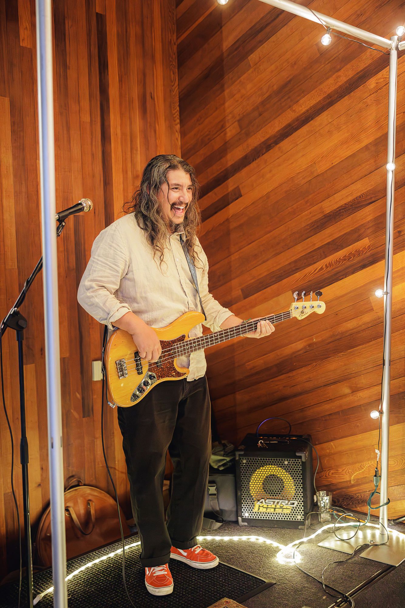 Musician plays electric bass guitar on wooden-paneled stage with dramatic lighting and performance equipment.