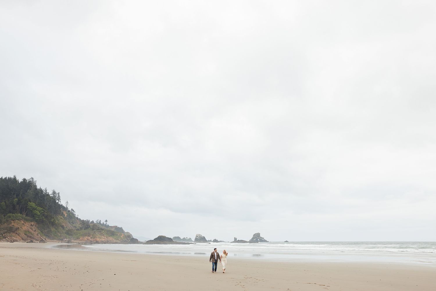 Couple walking hand in hand on a misty Oregon beach with dramatic cliffs and sea stacks in the background.