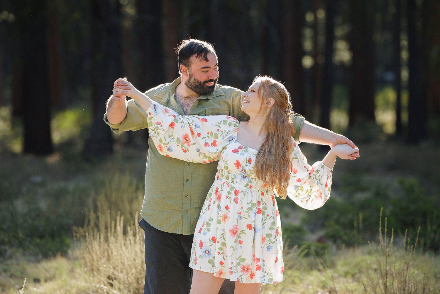 A couple dances joyfully in a sunlit forest, the woman wearing a floral dress and the man in a green shirt.