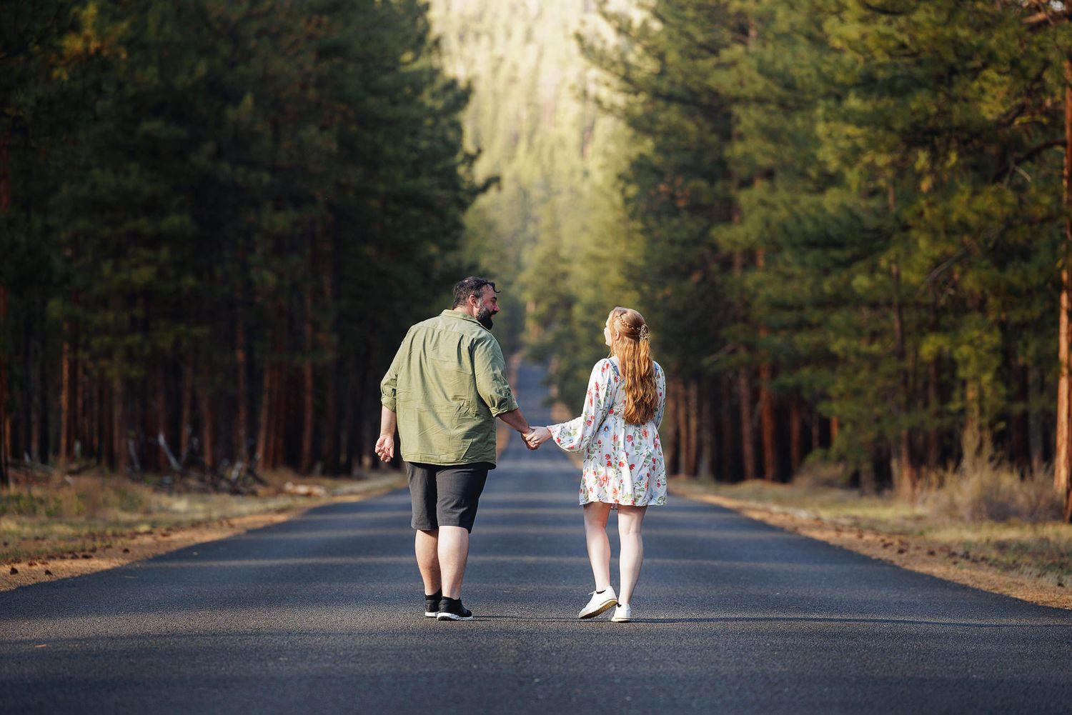 Adult and child walking hand in hand down a tree-lined road surrounded by tall pine forest.