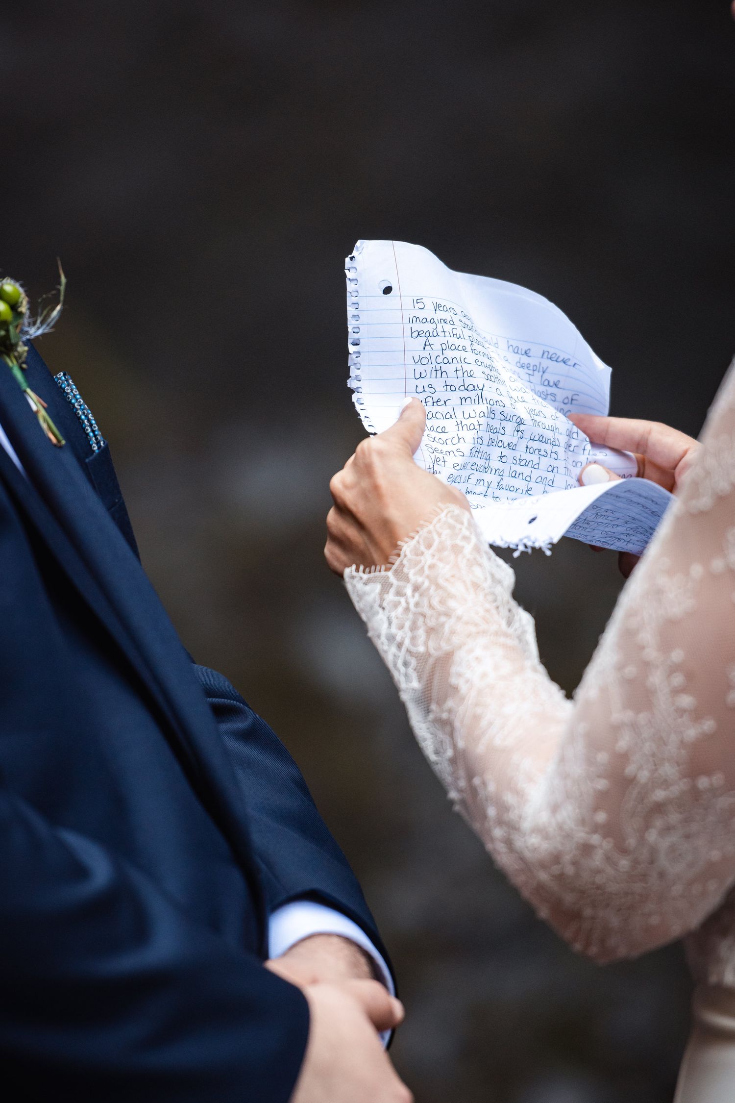 Hands hold a handwritten wedding vow note during a ceremony with the groom in a navy suit and bride in lace dress.