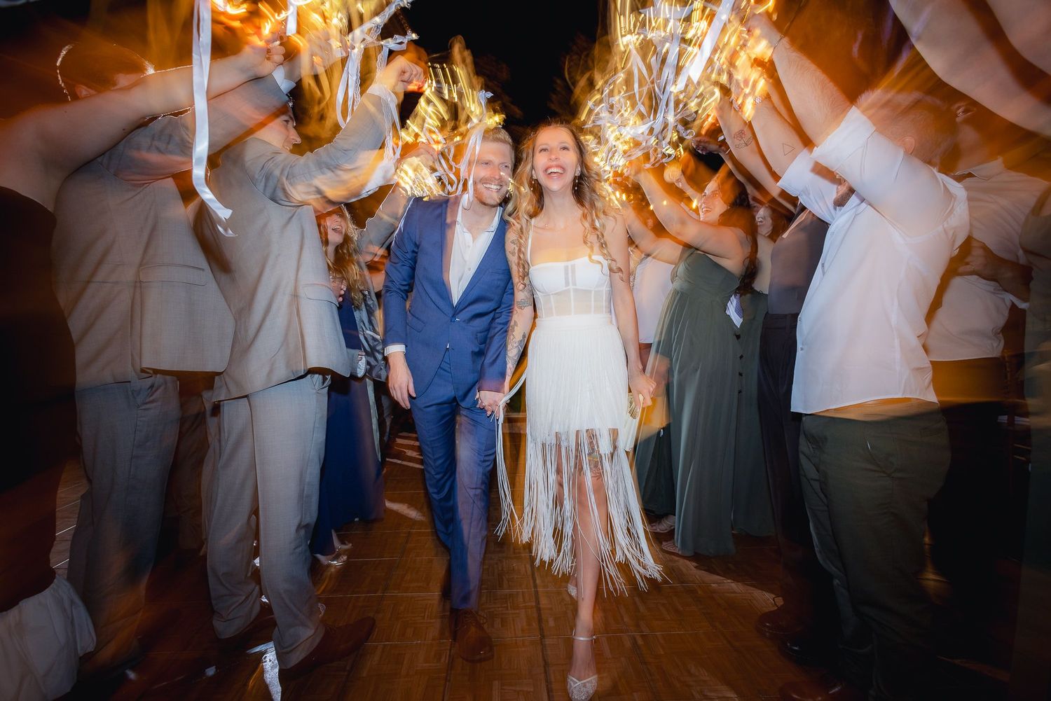 A couple walks through a sparkler-lined path surrounded by guests during their evening wedding celebration.