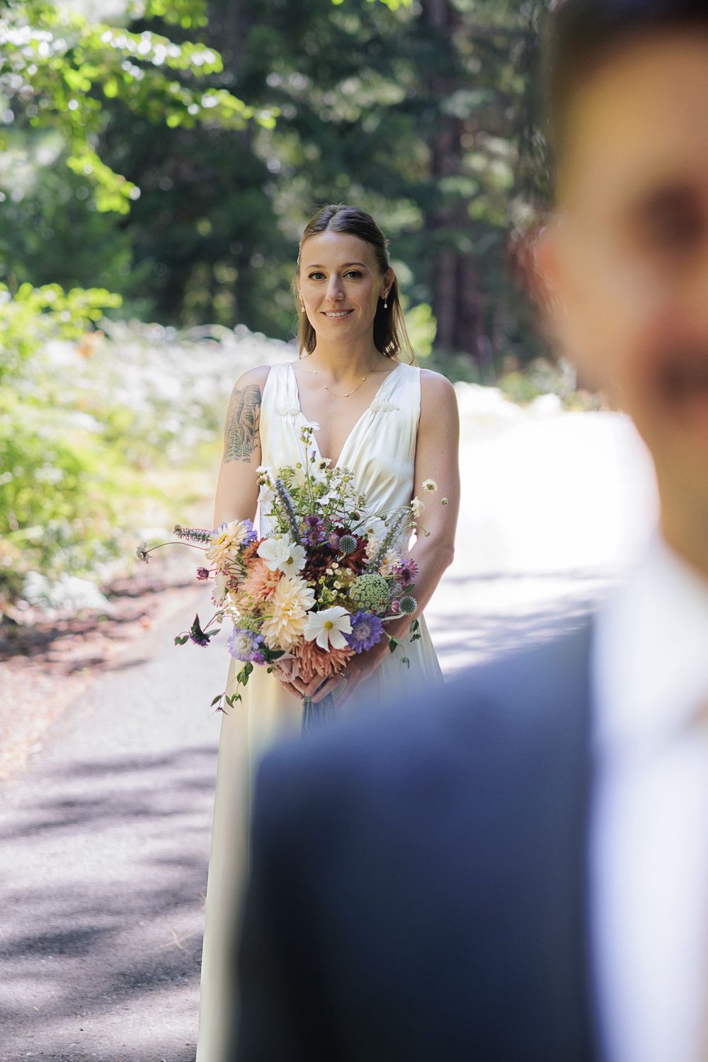 A person in a white wedding dress holds a beautiful wildflower bouquet while standing on a sunlit forest path.