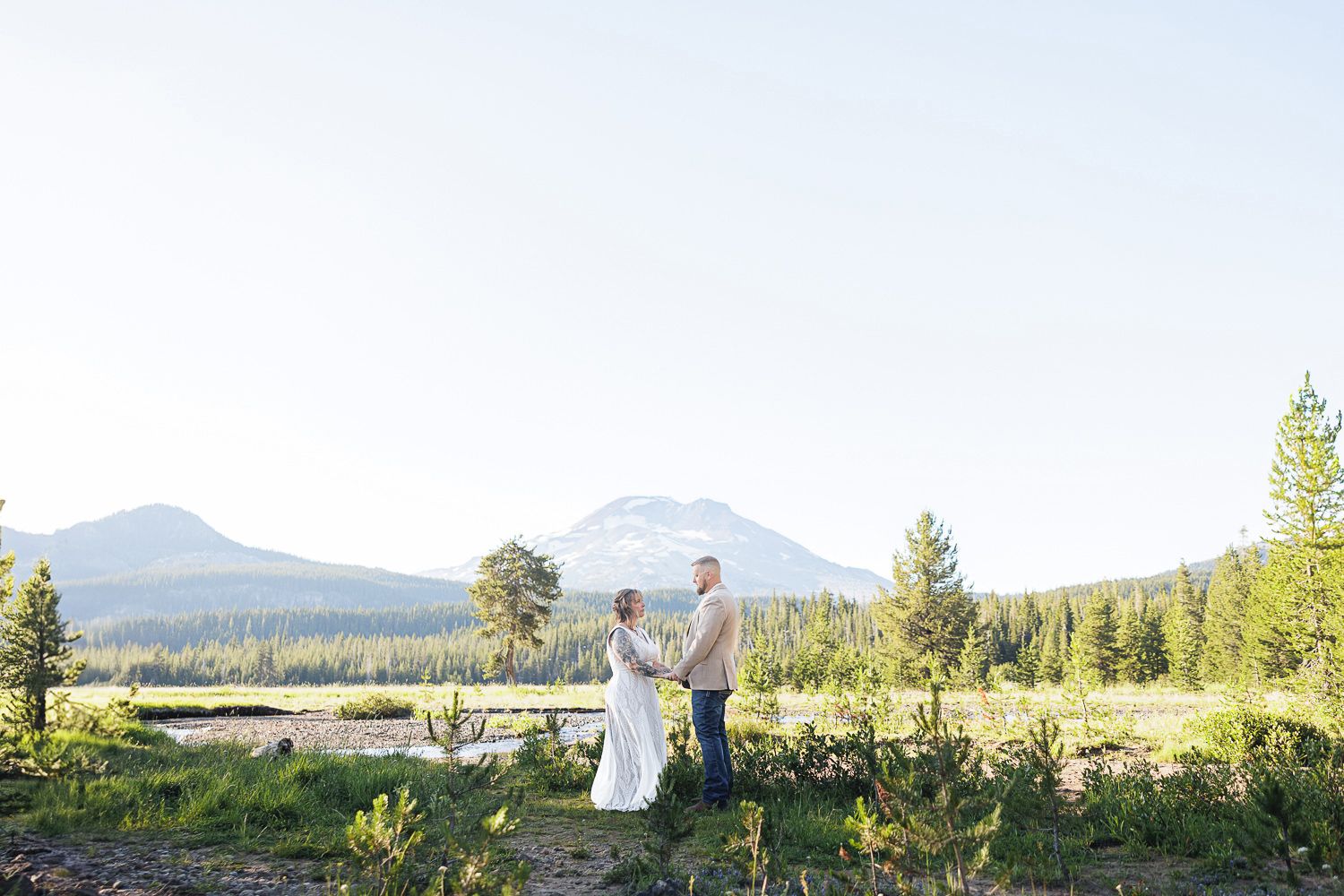 Two people celebrate in a scenic mountain meadow surrounded by evergreen trees and distant peaks at sunset.
