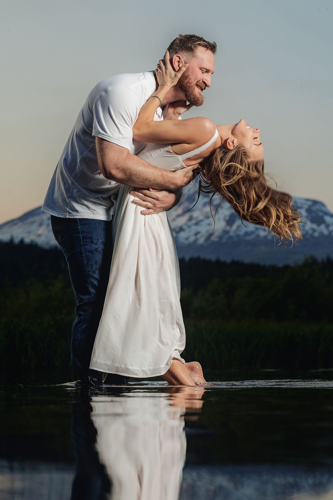 Couple in casual attire shares a romantic moment while standing in shallow water with mountain landscape backdrop.