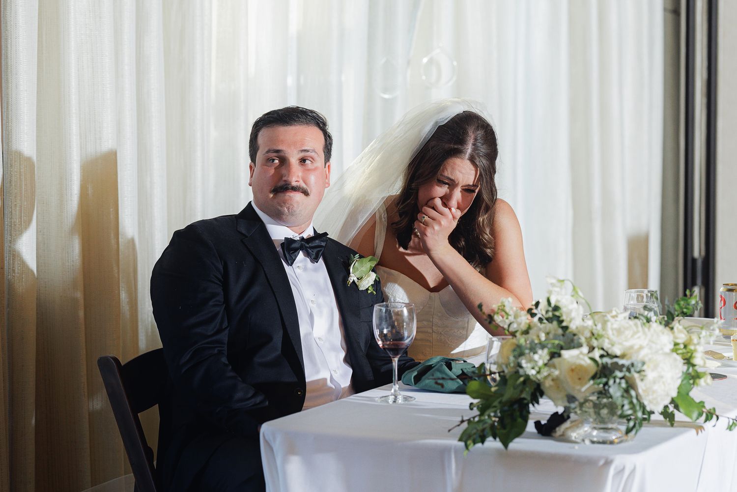 Bride and groom seated at wedding reception table, groom in tuxedo, bride in white gown laughing with floral centerpiece.