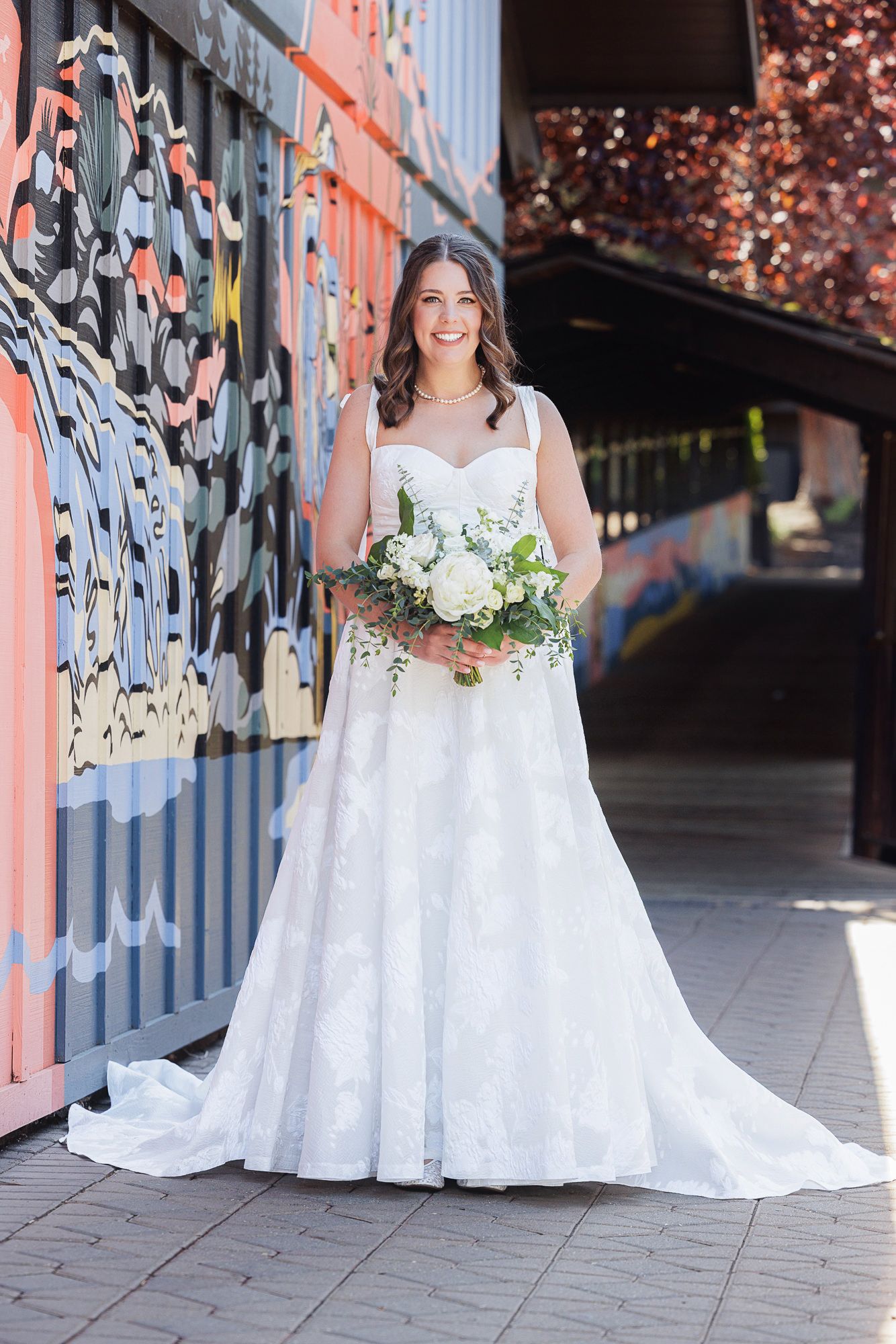 Smiling bride in lace A-line wedding gown holds white bouquet beside colorful graffiti wall outdoors.