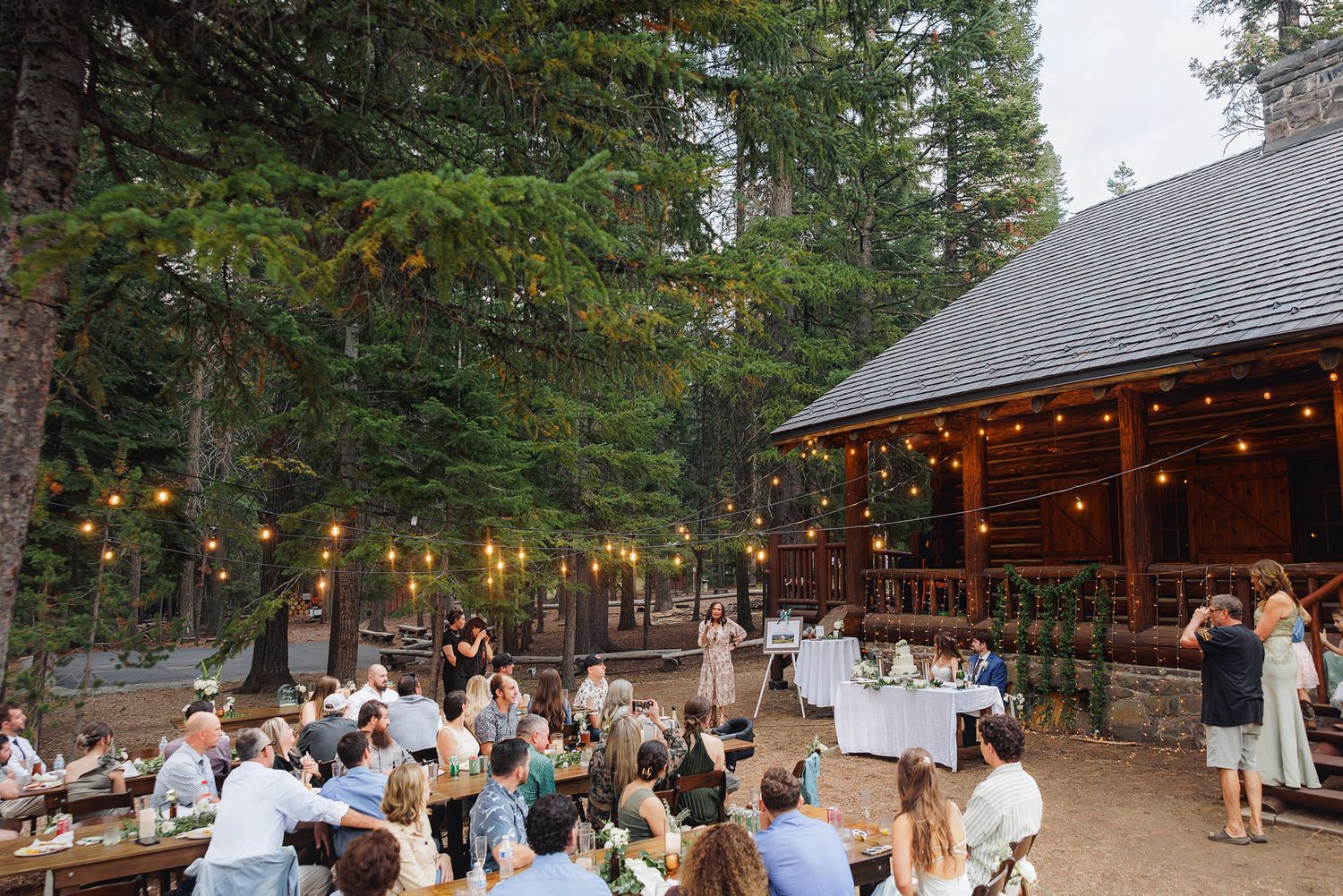 Outdoor wedding reception at a rustic mountain lodge with string lights and forest backdrop in the evening.