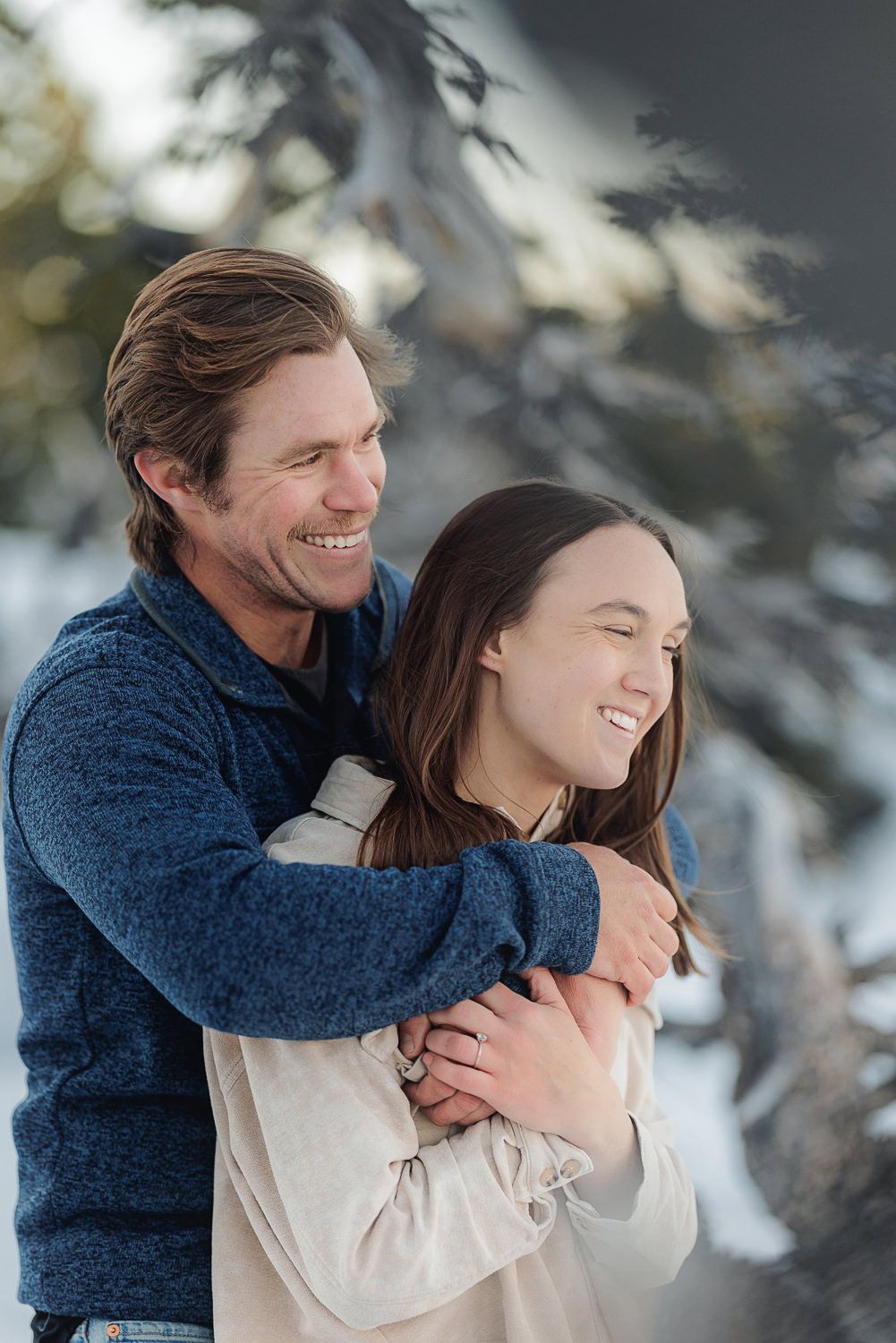 A couple shares playful moments and laughter in a snowy winter setting surrounded by evergreen trees.