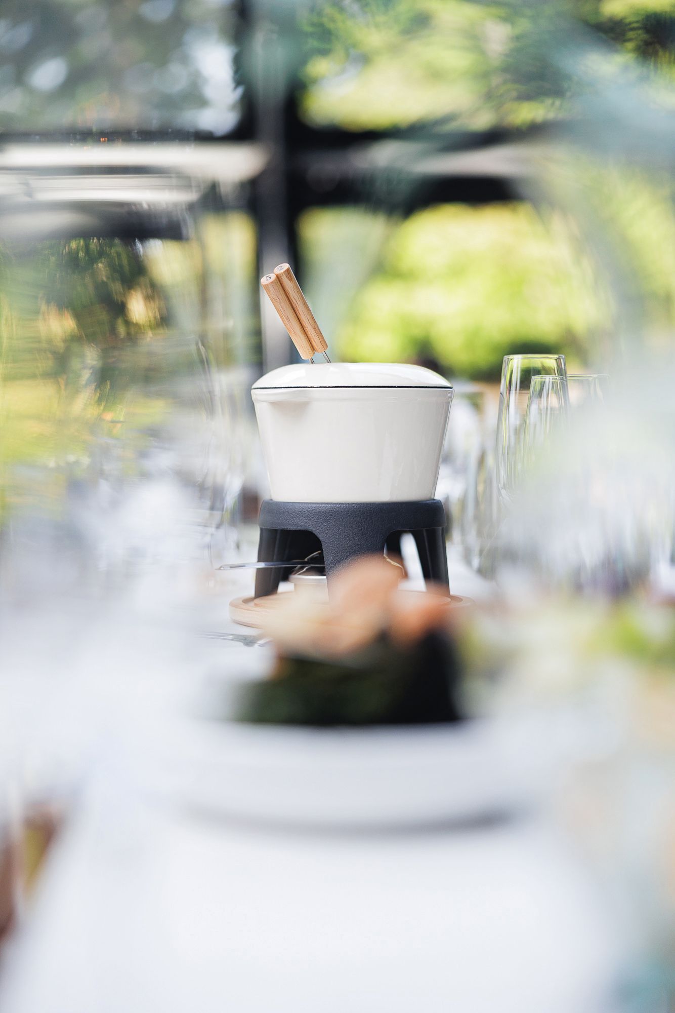 White fondue pot on a stand set on an elegant dining table with glassware, surrounded by lush greenery.