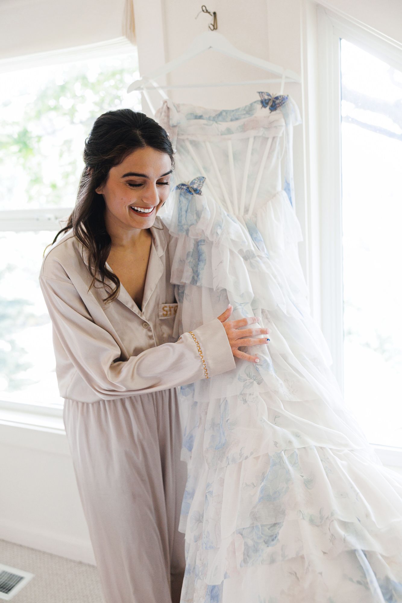 Bride in gray pajamas smiling while holding a floral blue and white wedding dress hanging by a window.