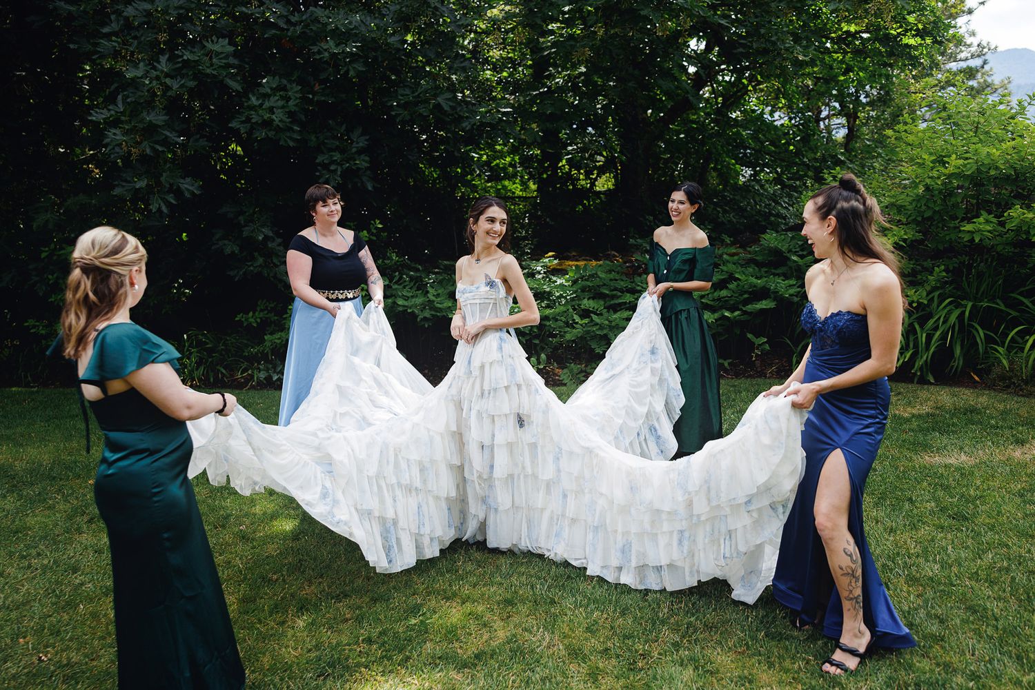 Bride in white gown with bridesmaids in green dresses holding train outdoors in garden setting.