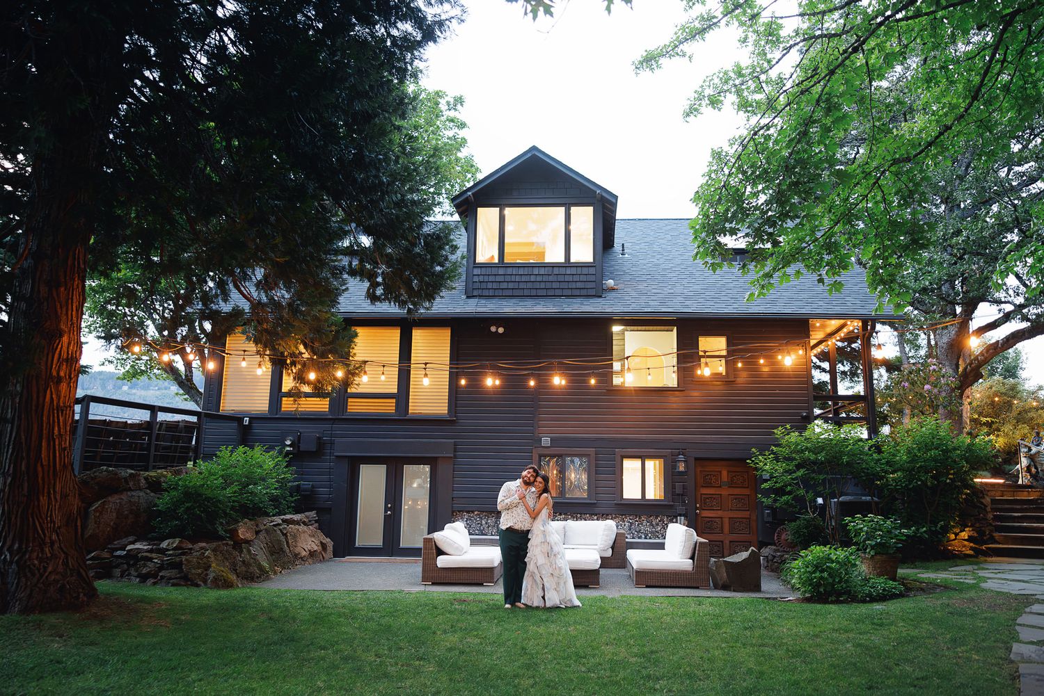 Couple embracing in wedding attire in front of a charming dark gray house with string lights at dusk.