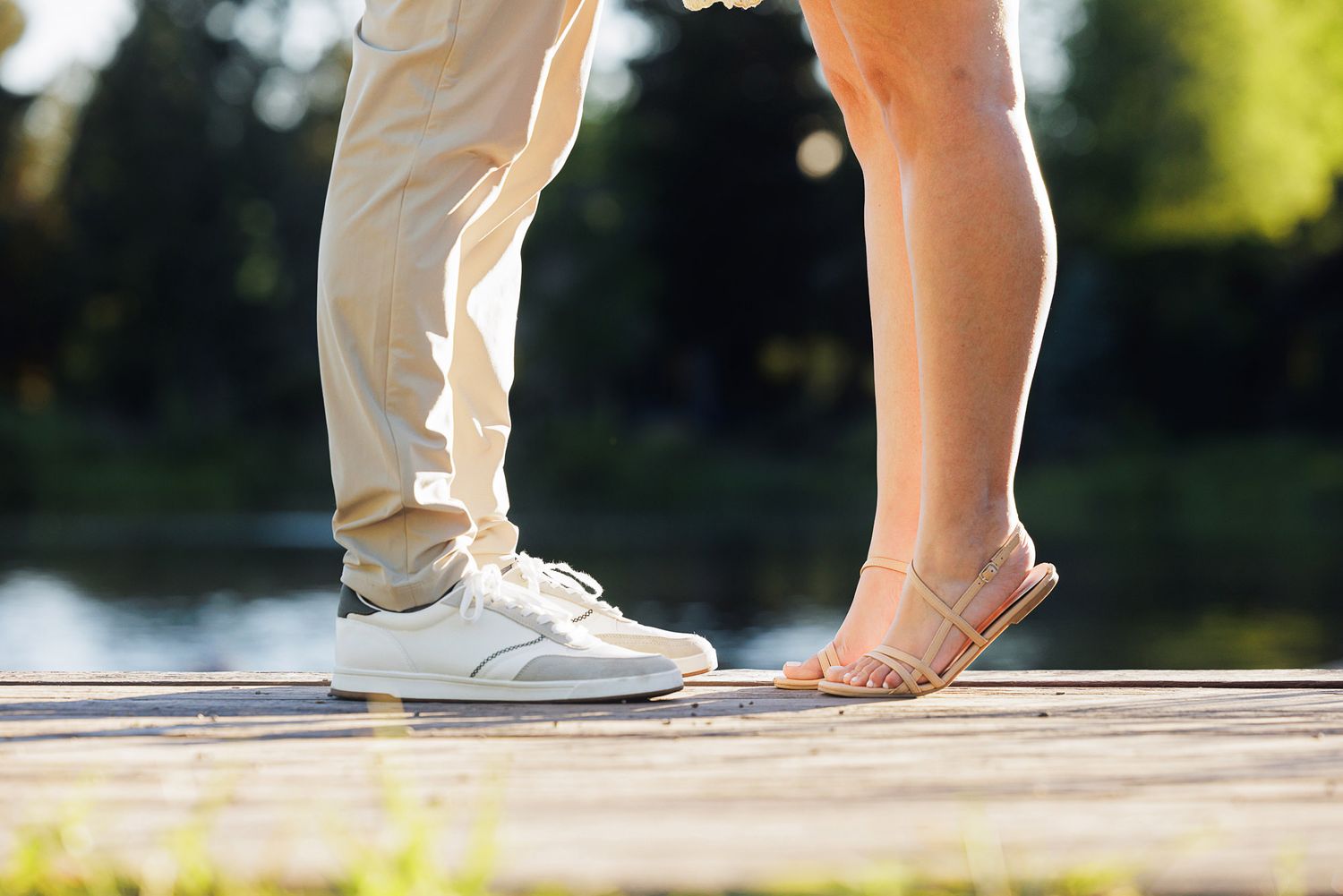 Close-up of a couple standing on a wooden dock, wearing casual shoes, sharing a romantic moment outdoors.