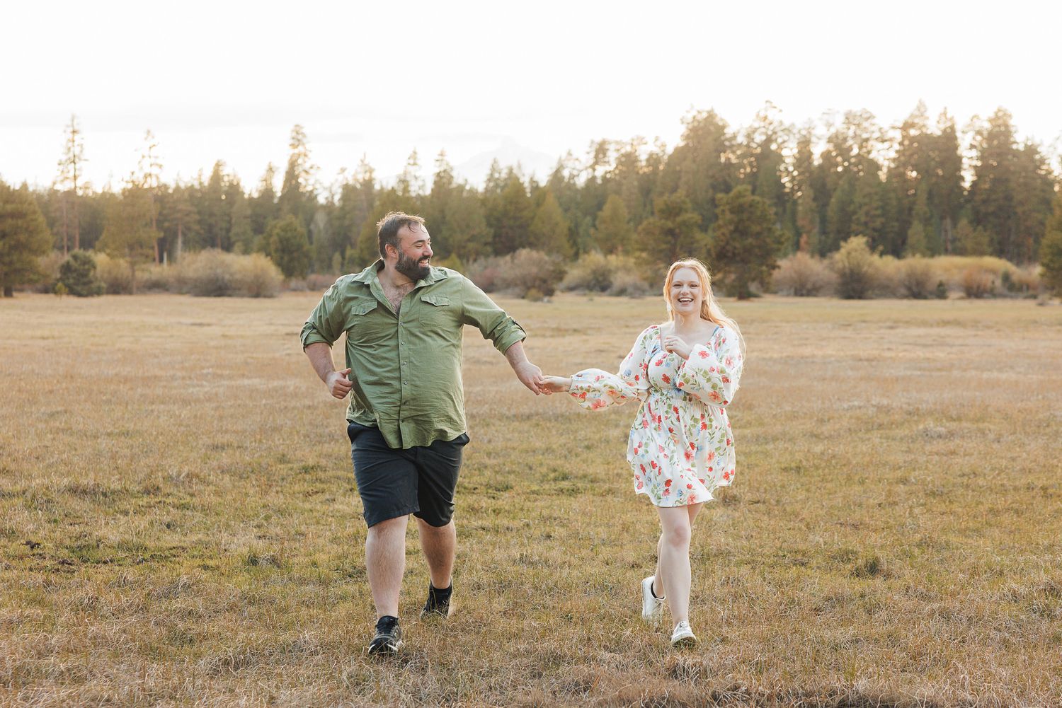 Happy couple holding hands and running through a scenic open field with trees in the background.
