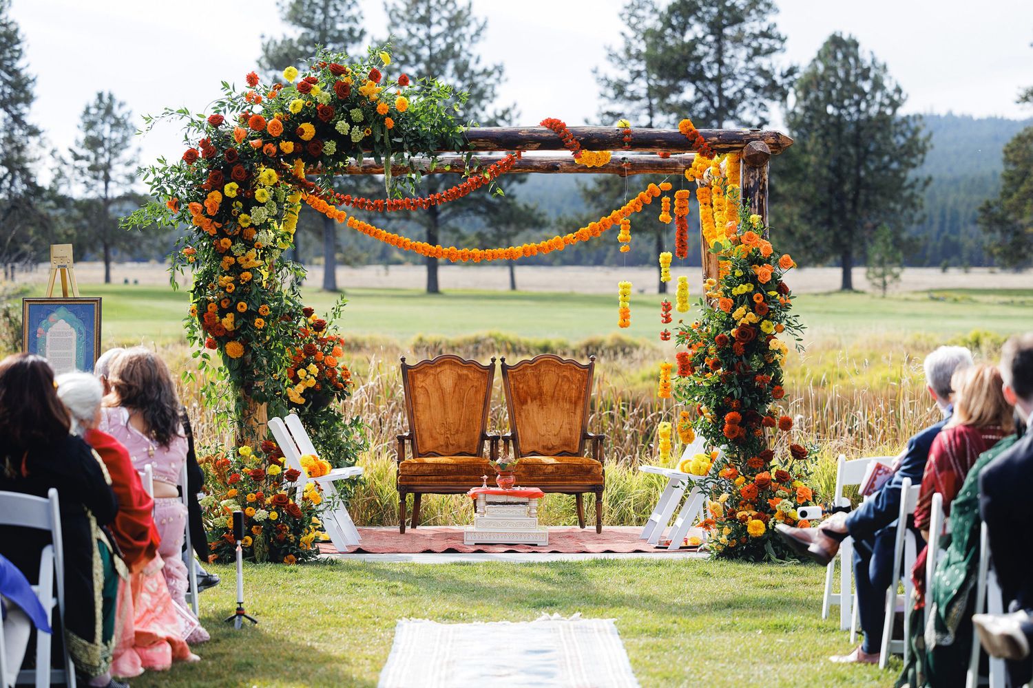 Outdoor Indian wedding ceremony with floral arch of orange and yellow blooms, wooden chairs, and white aisle on a golf course.