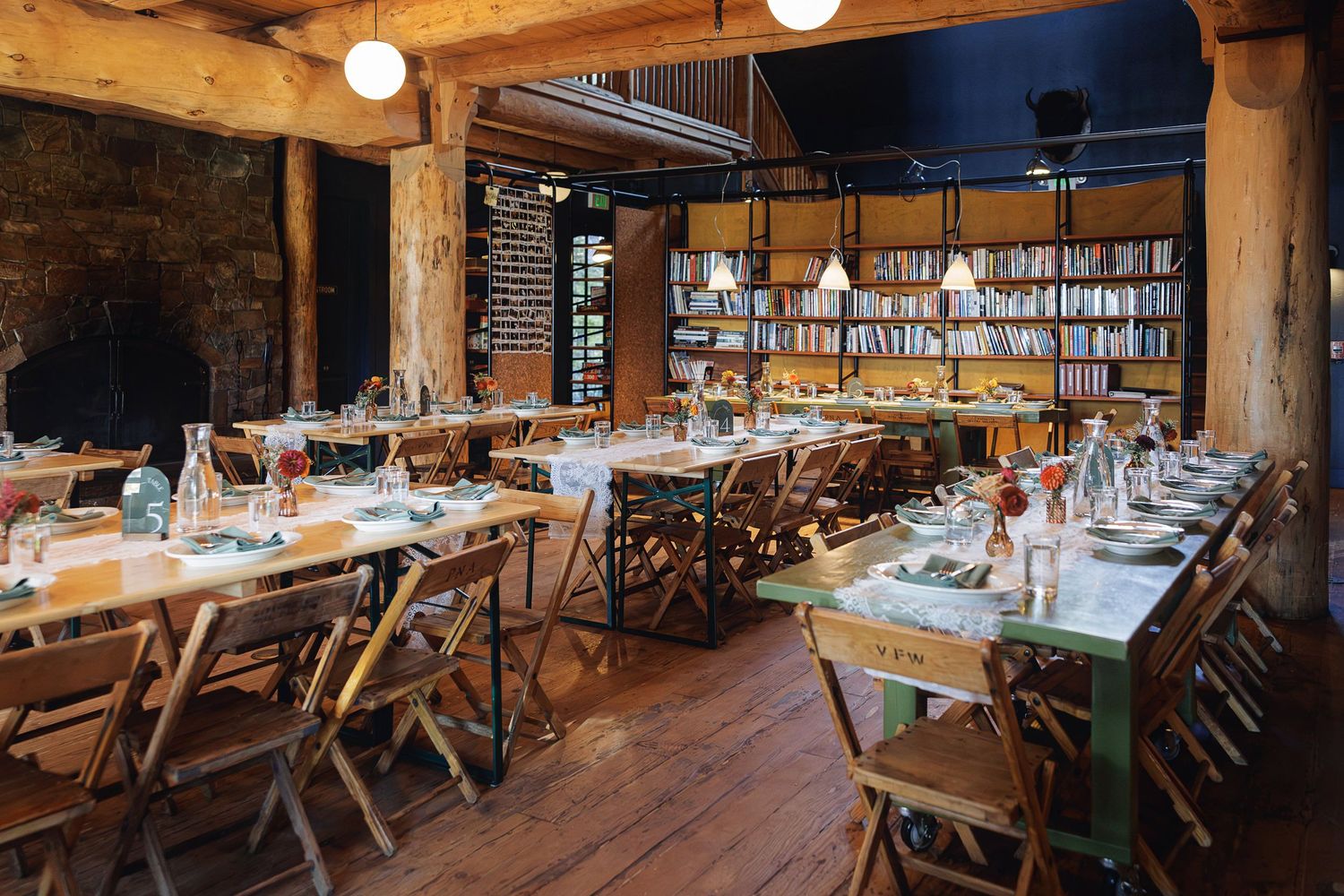Rustic restaurant dining room with wooden tables, folding chairs, globe lights, and floor-to-ceiling bookshelves along the back wall.