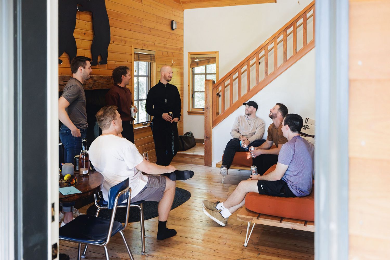 Group of men socializing in a cozy wooden cabin living room with stairs and natural light.