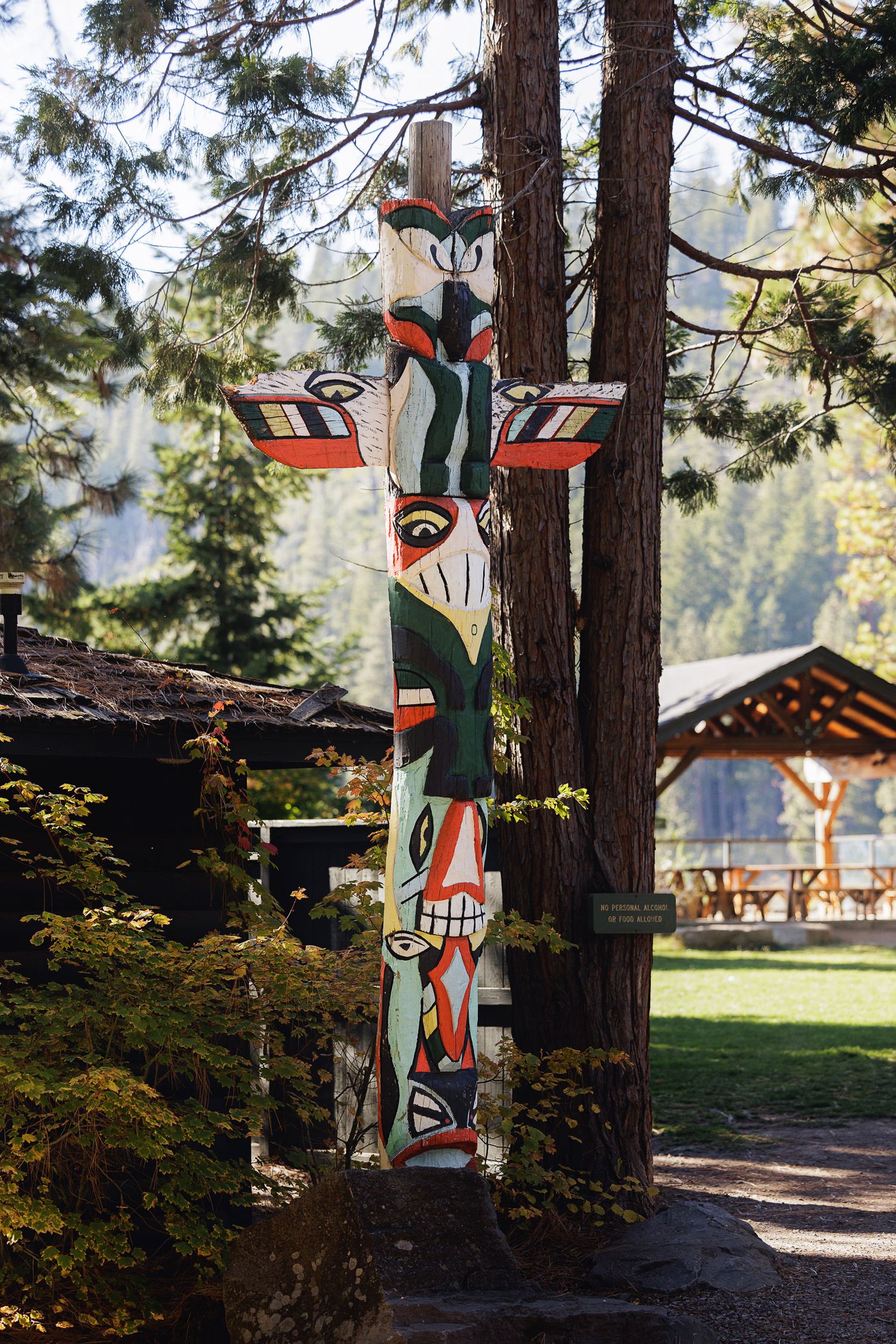 Colorful Native American totem pole standing tall among pine trees near a wooden pavilion in a forested park setting.