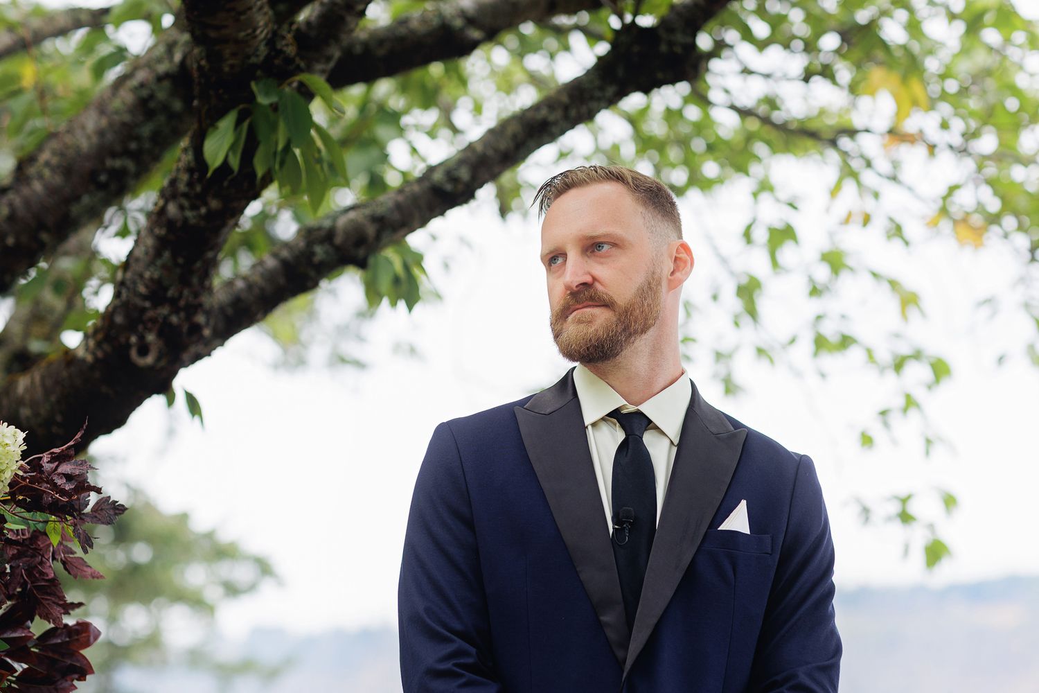 Groom in navy tuxedo with black lapels and white pocket square standing outdoors under a tree at a wedding.