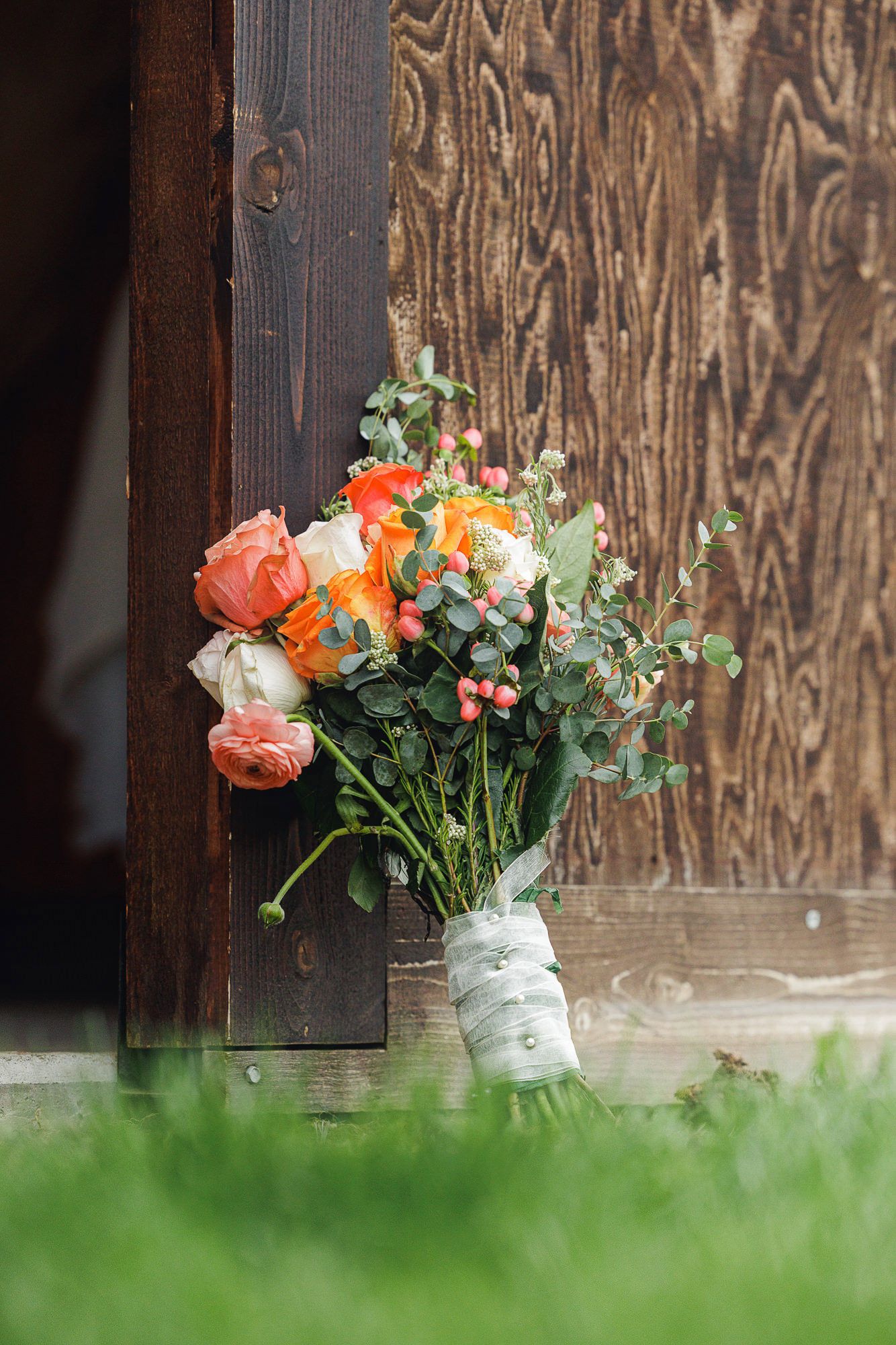 A rustic wedding bouquet of peach roses and eucalyptus tied with white ribbon, leaning against a dark wooden barn door.