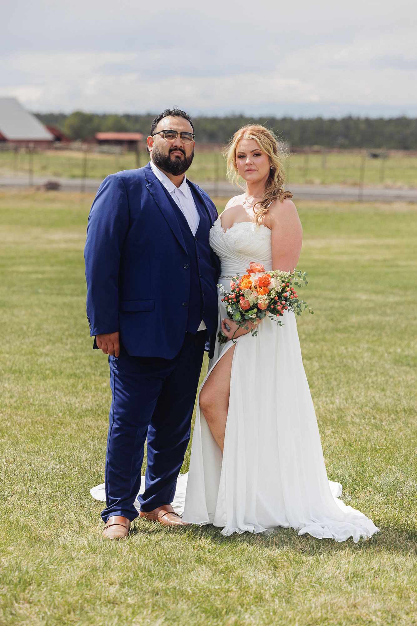 Bride in white gown holding orange bouquet stands with groom in navy suit on green lawn at outdoor farm wedding.