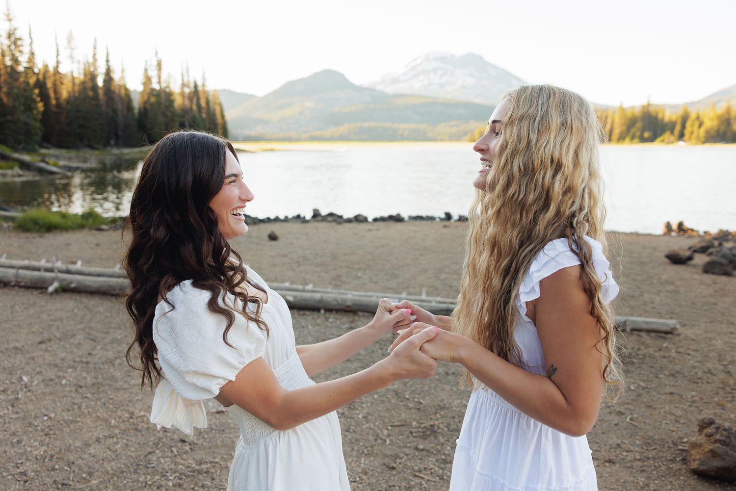 Two women in white dresses laugh and hold hands by a mountain lake surrounded by pine trees.