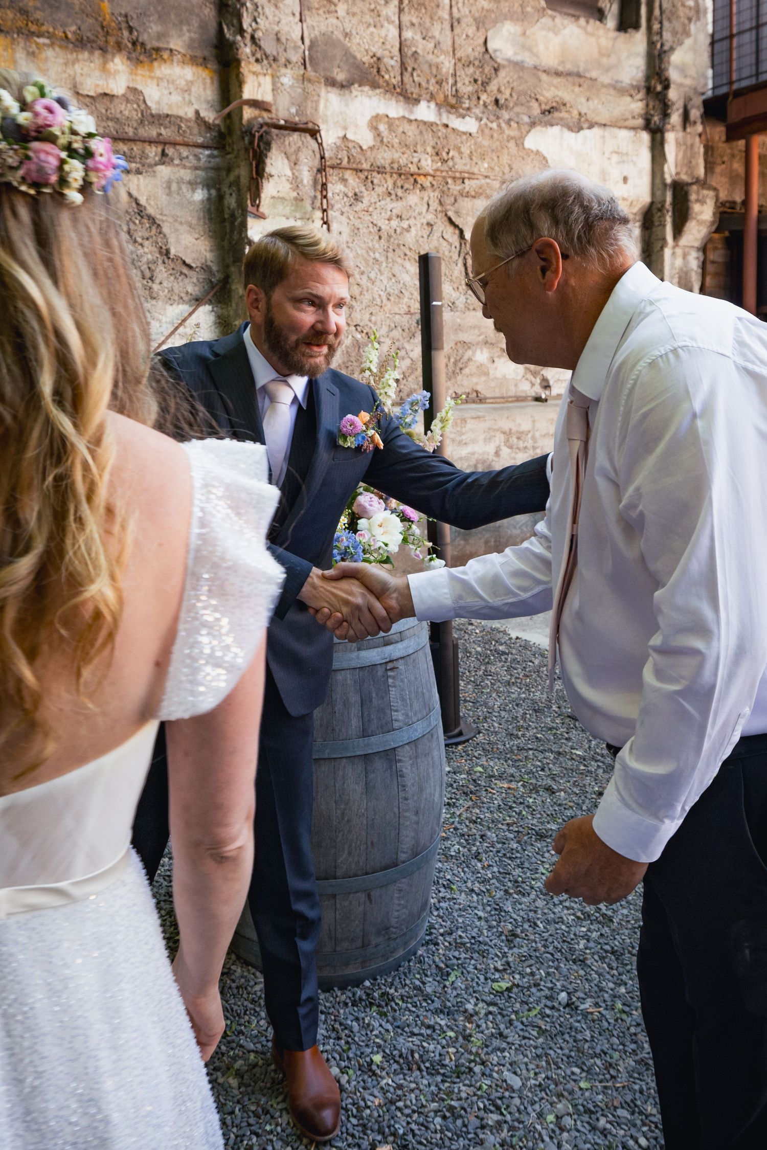 Wedding guests greet each other during outdoor reception by rustic stone wall and wine barrel.