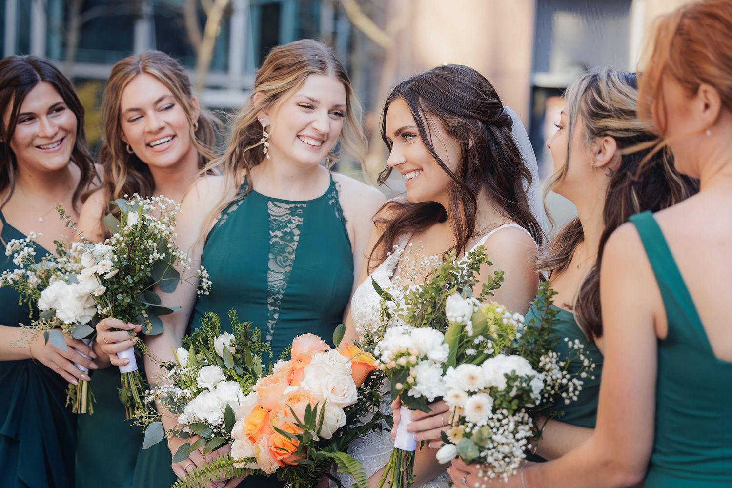 Bridesmaids in matching emerald green dresses hold white and peach floral bouquets while laughing together.