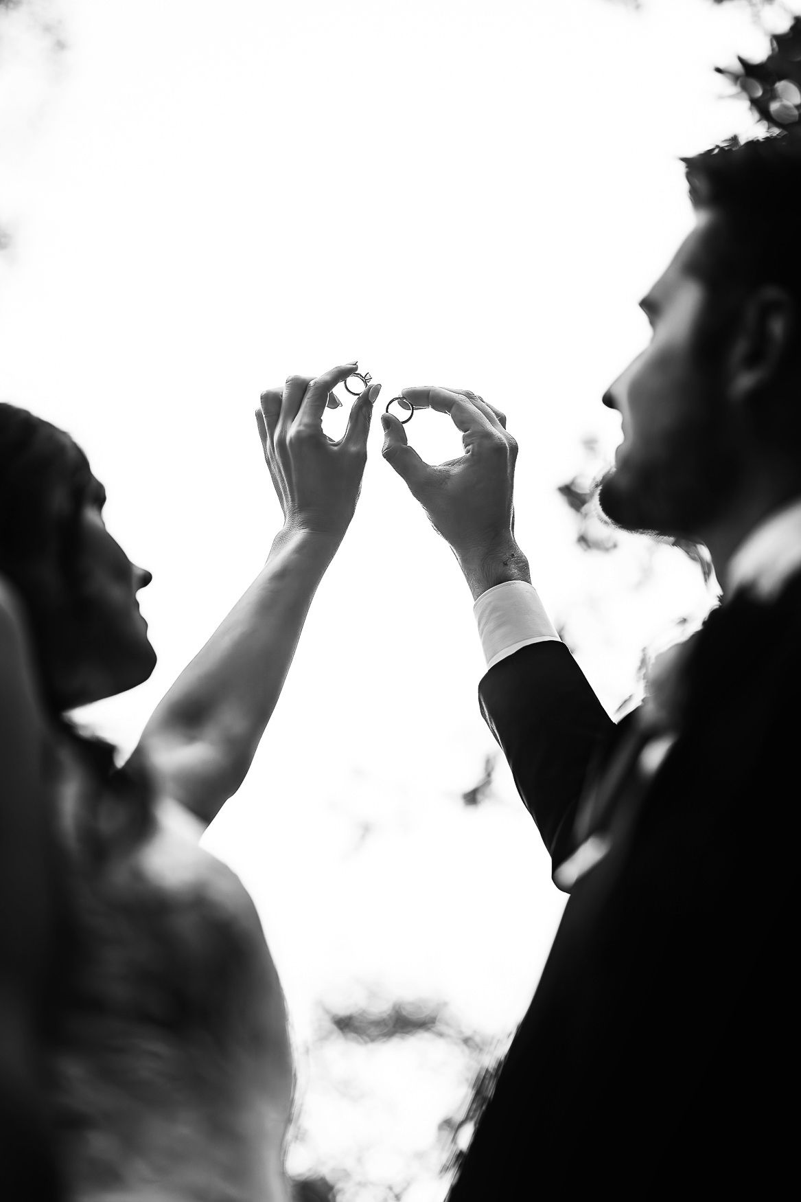 Black and white silhouetted figures reaching up to make heart shape with hands against bright background.