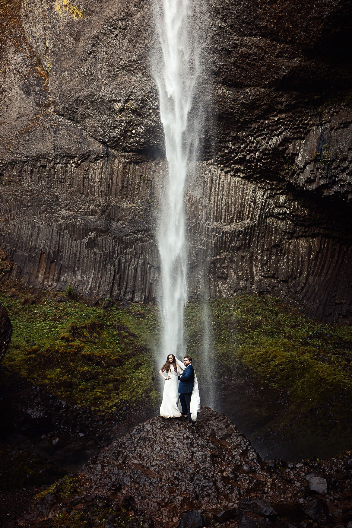 A couple poses beneath a dramatic tall waterfall against rocky cliffs.