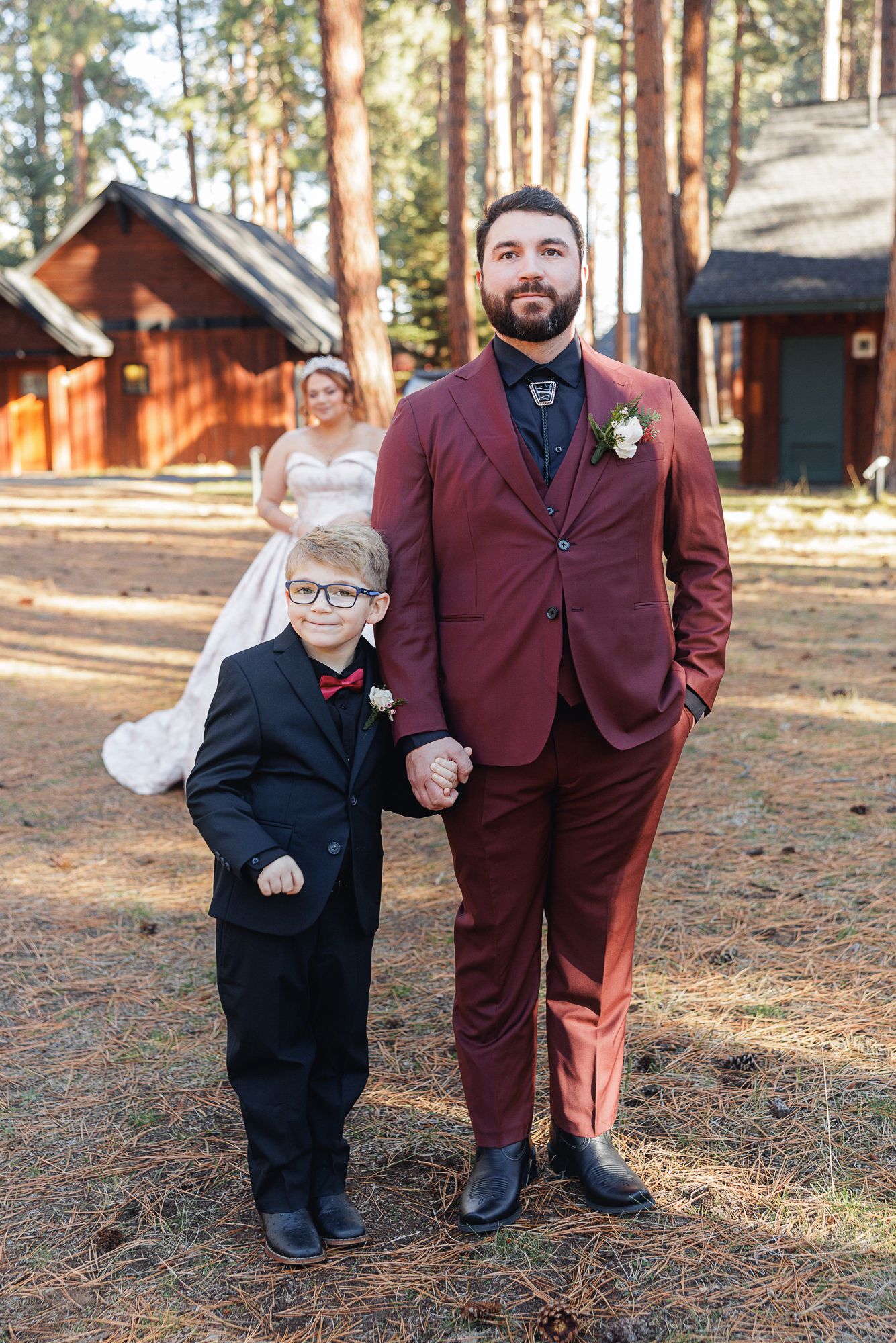 A young boy in a black suit and adult in burgundy suit stand together on forest ground surrounded by tall pine trees.