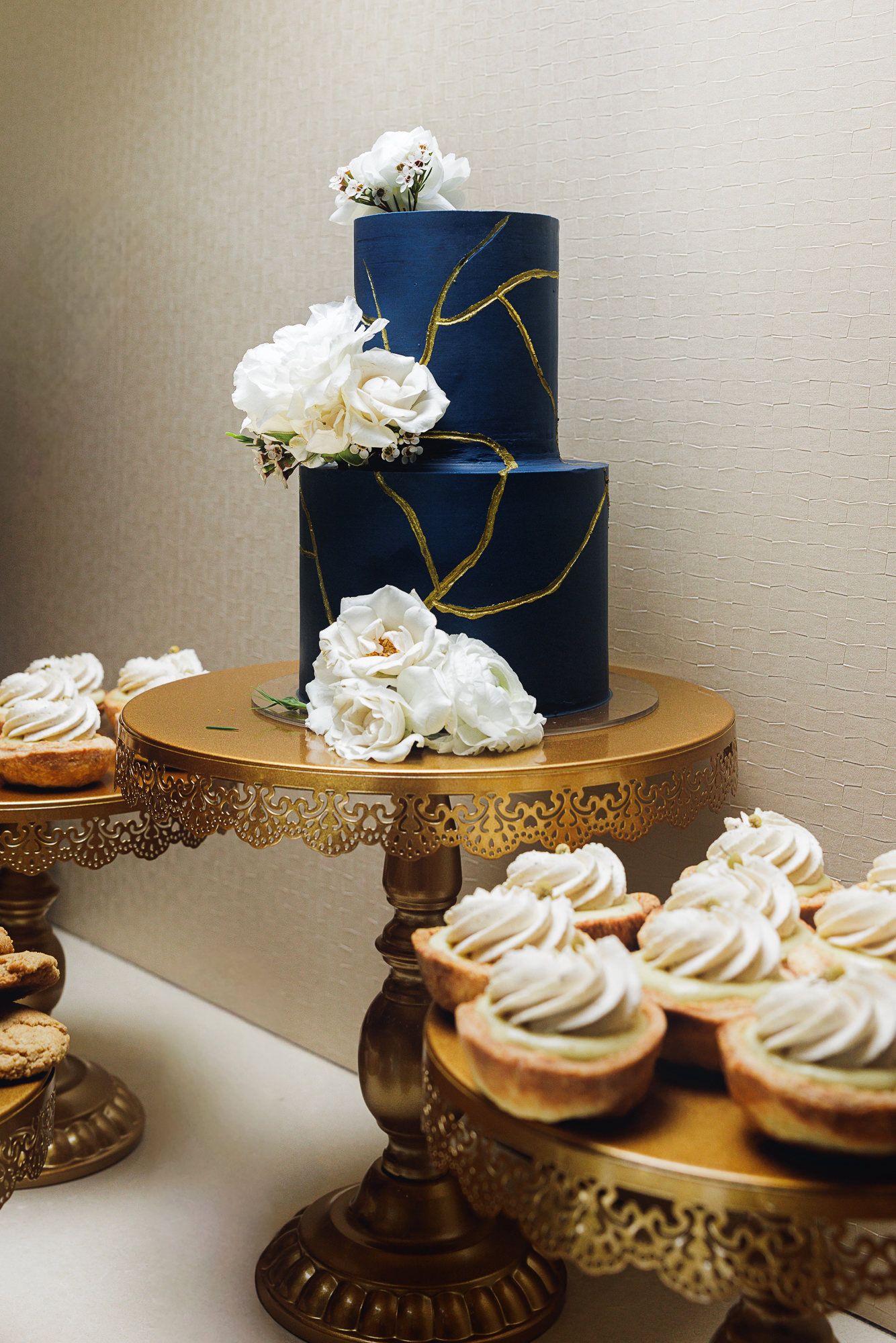 Navy blue wedding cake with gold accents and white roses displayed alongside cupcakes on ornate gold cake stands.