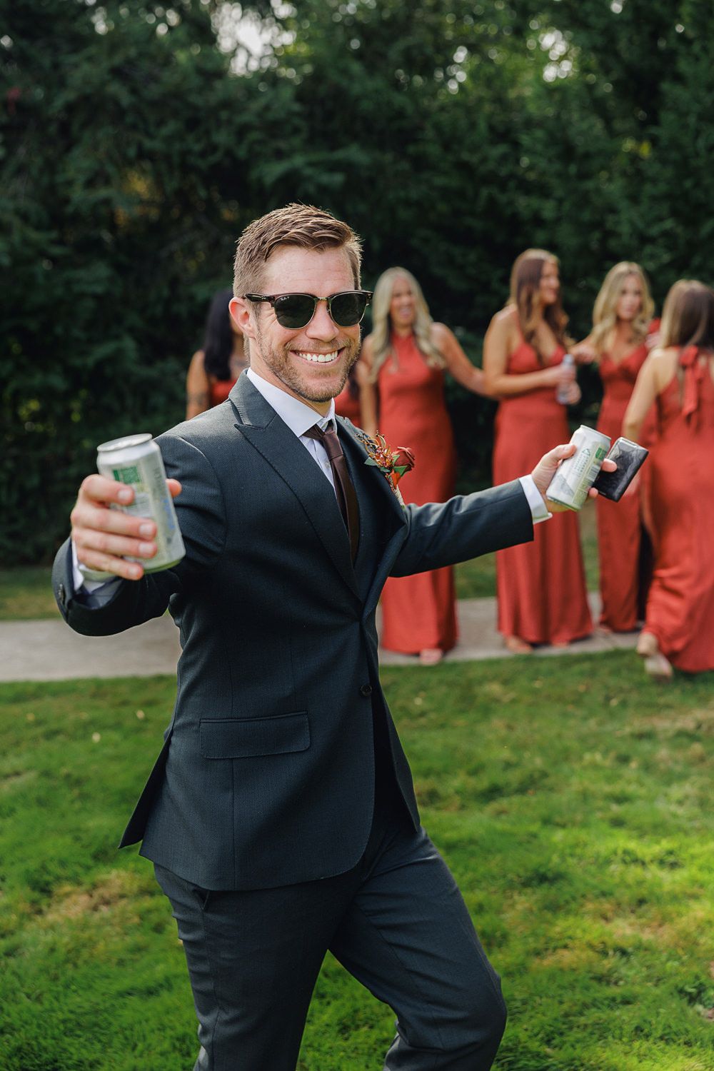 A groomsman in a dark suit and sunglasses holds up drinks while celebrating at an outdoor wedding with bridesmaids in red.