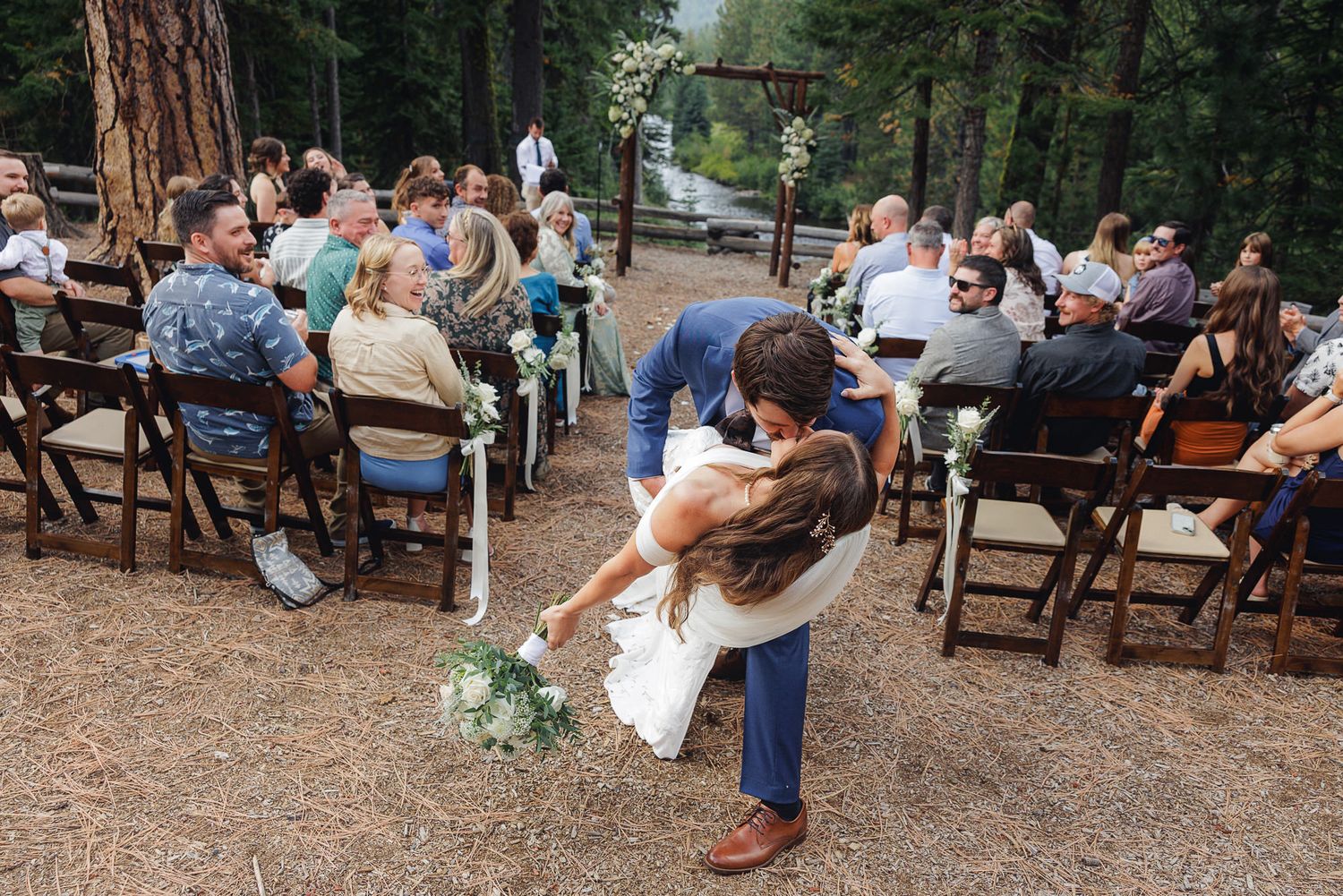 Wedding guests gather outdoors at rustic wooden benches in a forest setting during a casual ceremony celebration.