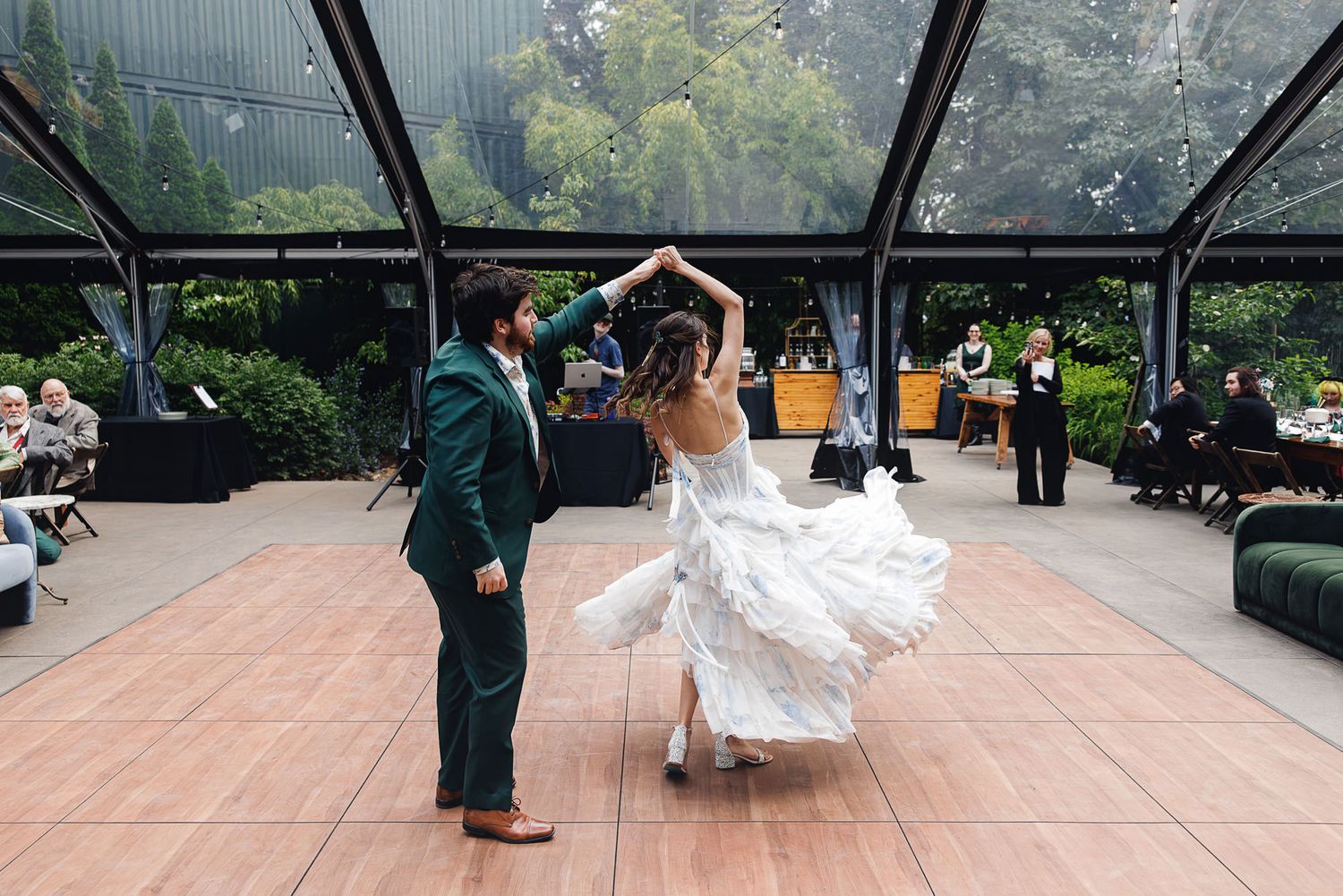 Bride and groom share their first dance under a glass pavilion surrounded by lush greenery at an elegant outdoor wedding.