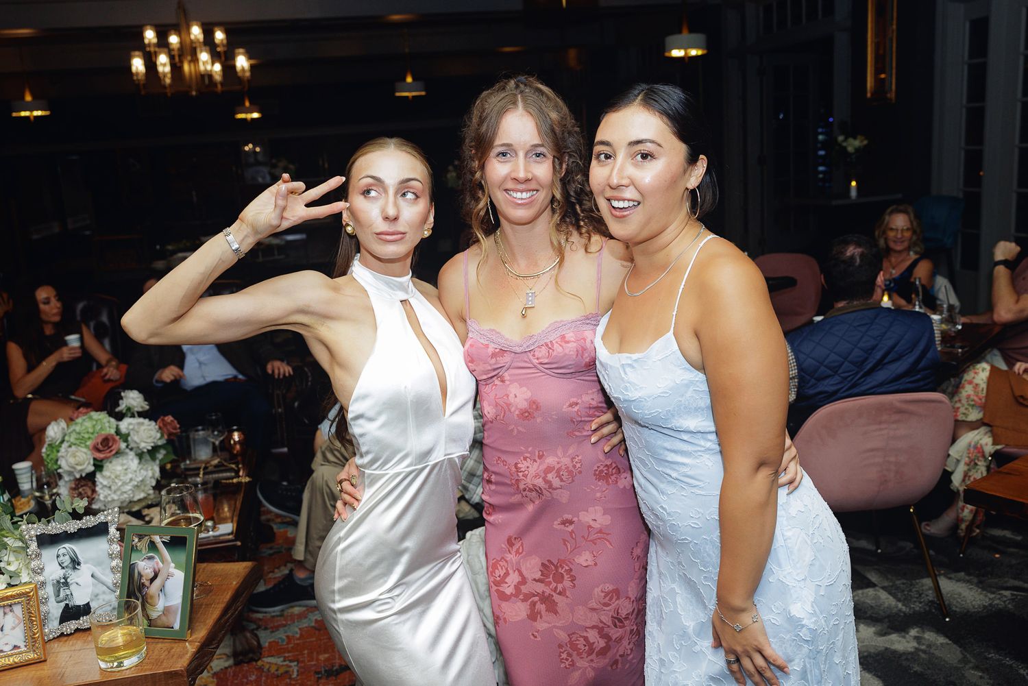 Three women in elegant dresses posing together at a formal event, one making a peace sign.