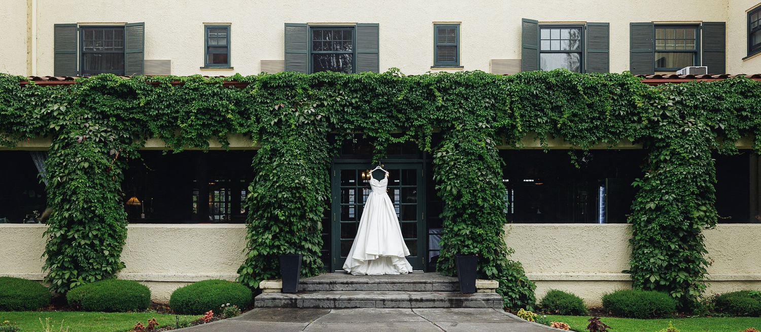 Bride in white wedding gown posing at ivy-covered elegant venue entrance with lush green walls and manicured hedges.