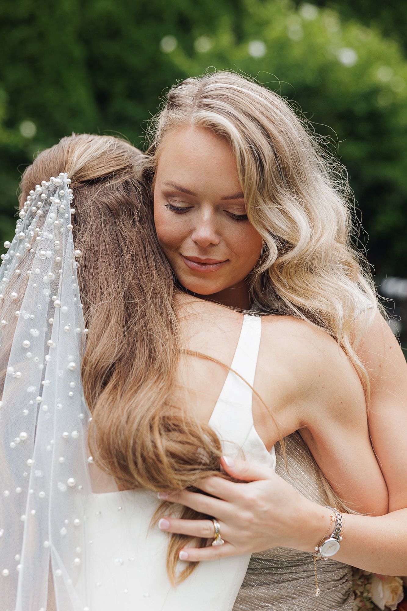 Bride in pearl veil embracing bridesmaid in emotional wedding hug outdoors with green trees background.