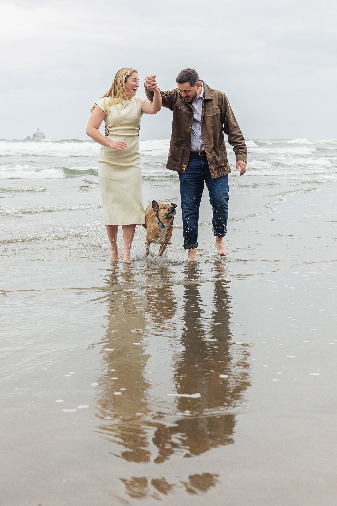 Couple dancing barefoot on a sandy beach with their French Bulldog in shallow ocean waves on a cloudy day.