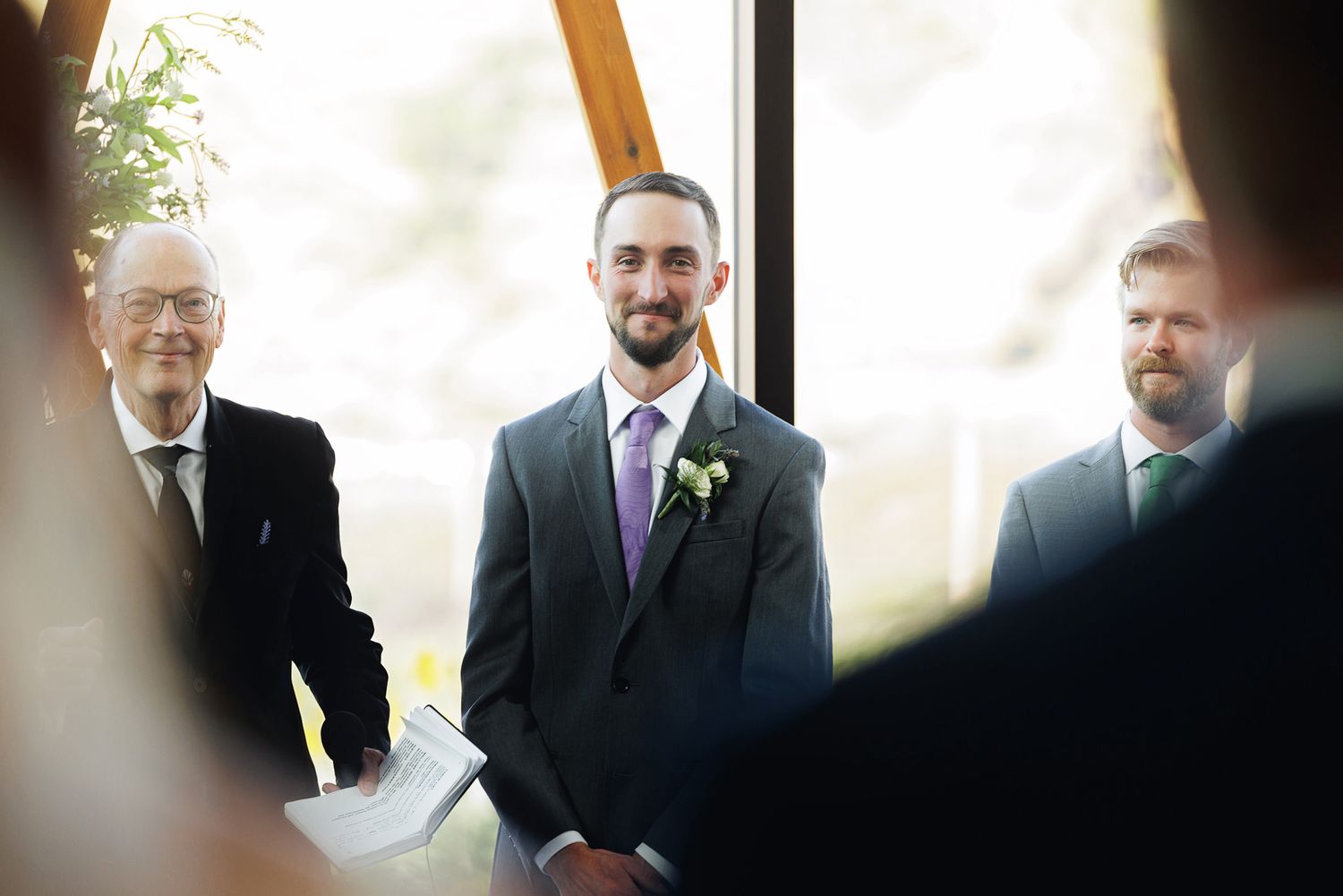 Groom smiling at the altar in a dark suit with purple tie and boutonniere during an indoor wedding ceremony.