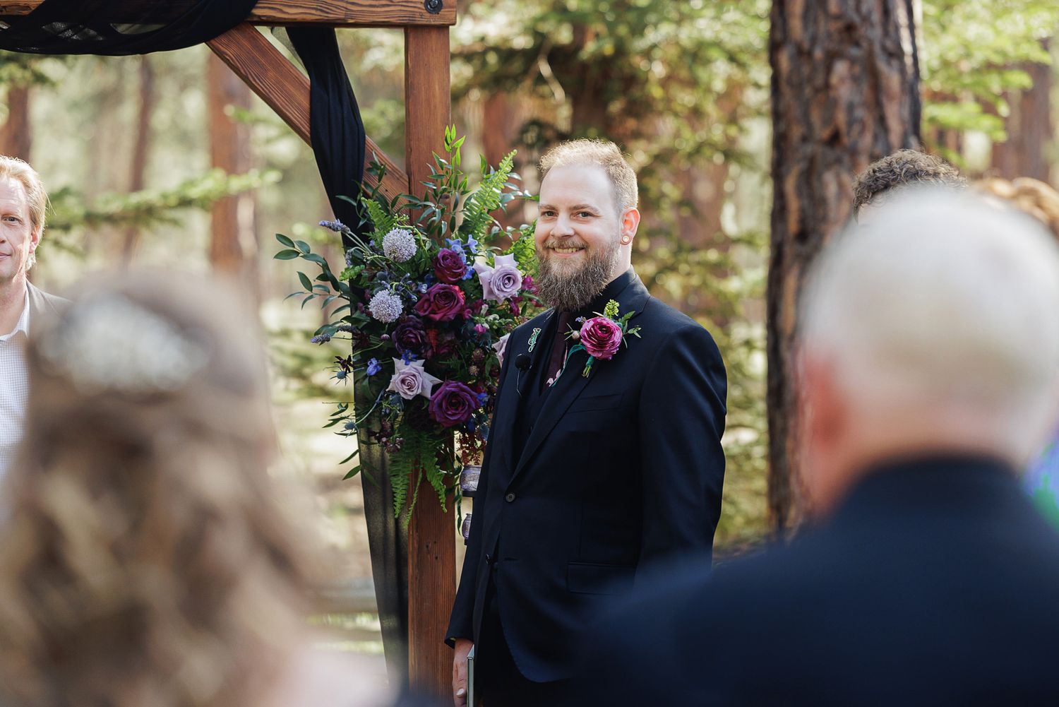 Groom in black suit smiling at altar during outdoor woodland wedding ceremony with purple floral arrangements.