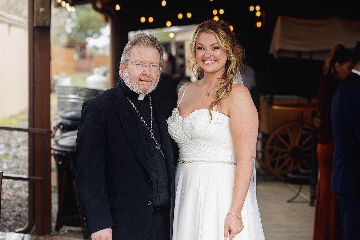 Bride in white strapless gown posing with a priest at an outdoor wedding venue with string lights.