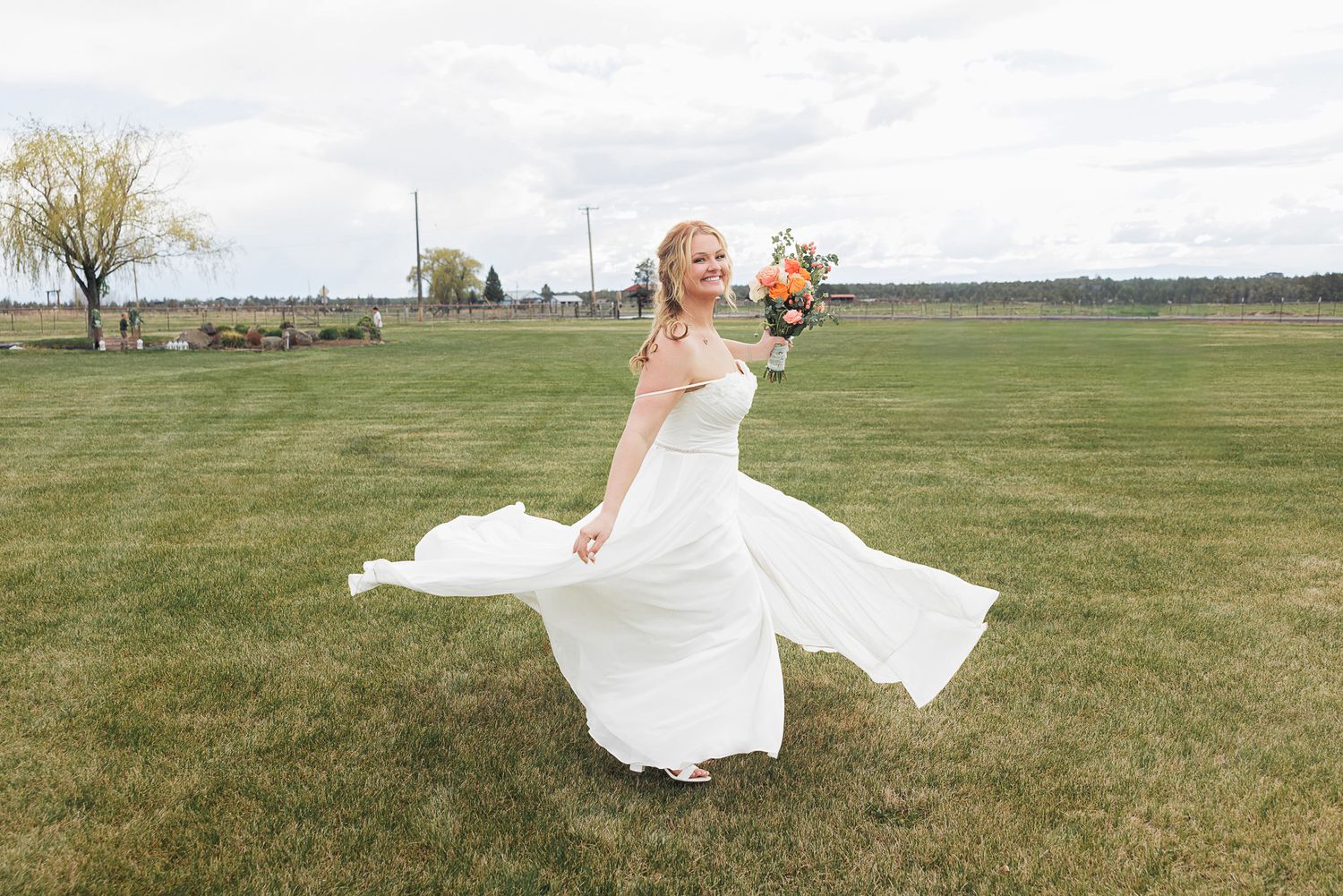 Bride in white gown twirling on green lawn holding colorful bouquet on her wedding day.