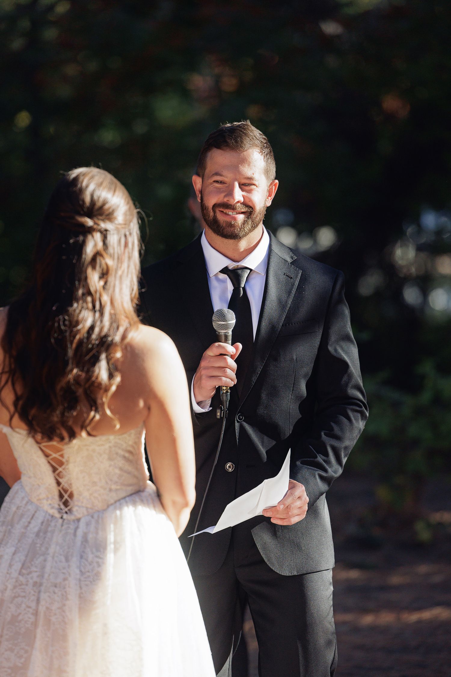 Groom smiling joyfully while reading wedding vows into a microphone, holding paper, wearing dark suit with black tie outdoors.