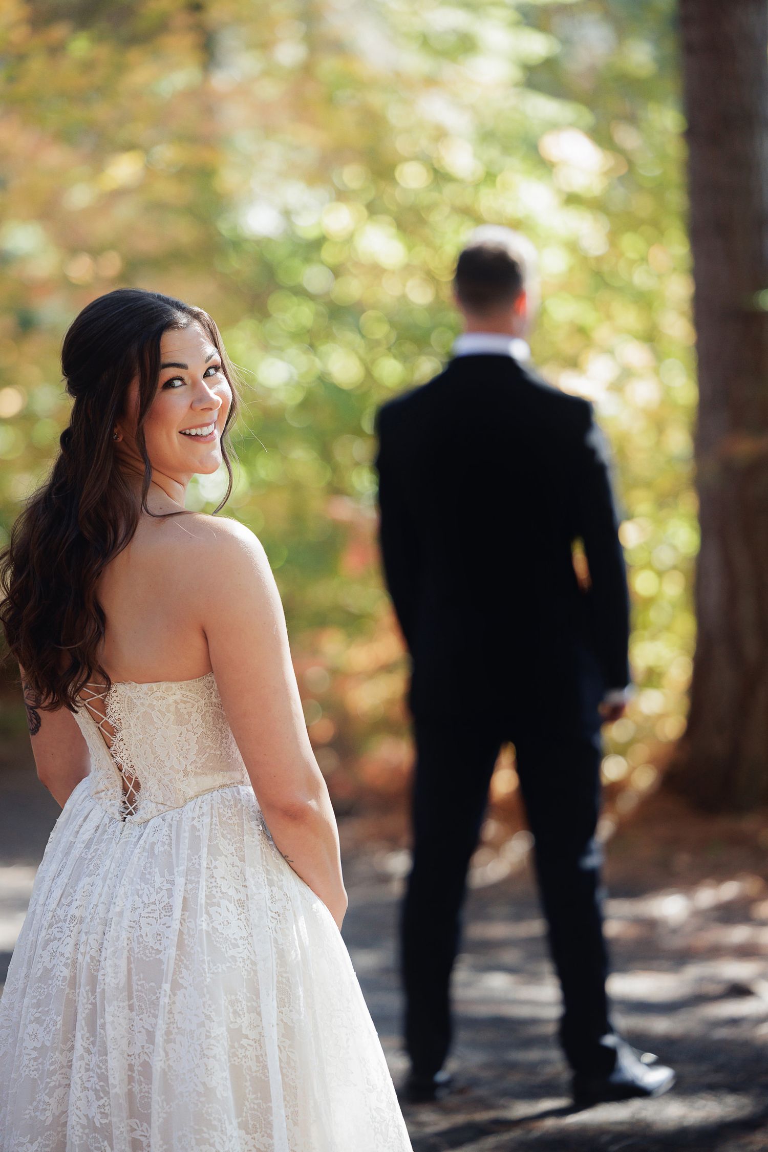 Bride in strapless lace wedding gown smiles over shoulder as groom stands in background outdoors.