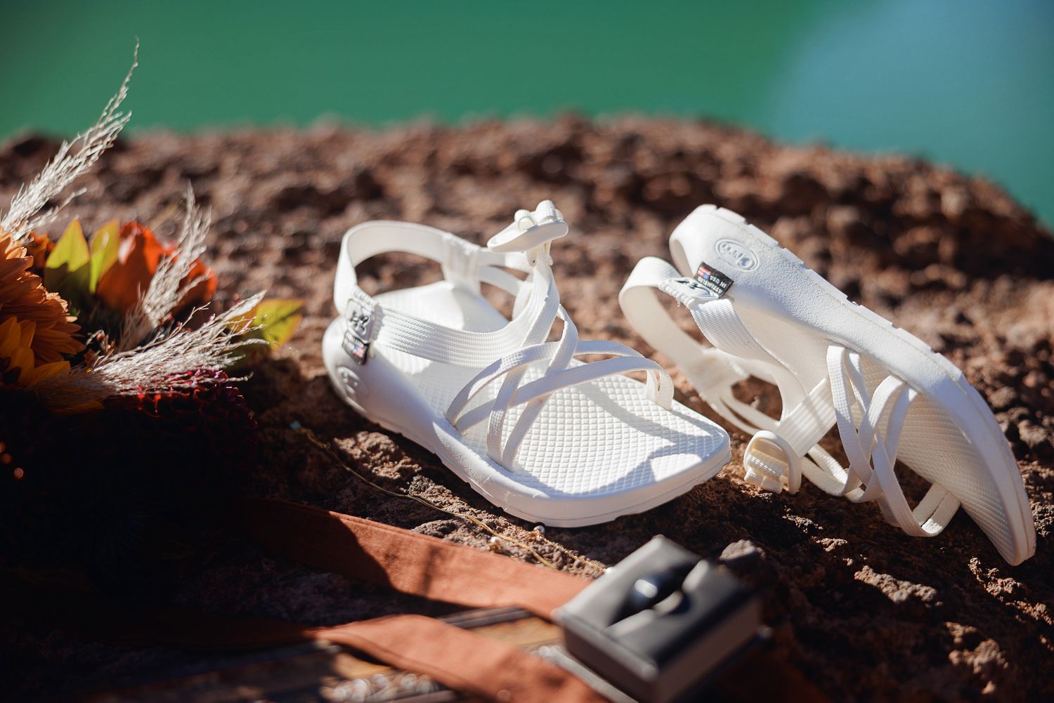 White strappy sport sandals rest on rust-colored rocks beside autumn flowers and dried grass, with a small ring box nearby.