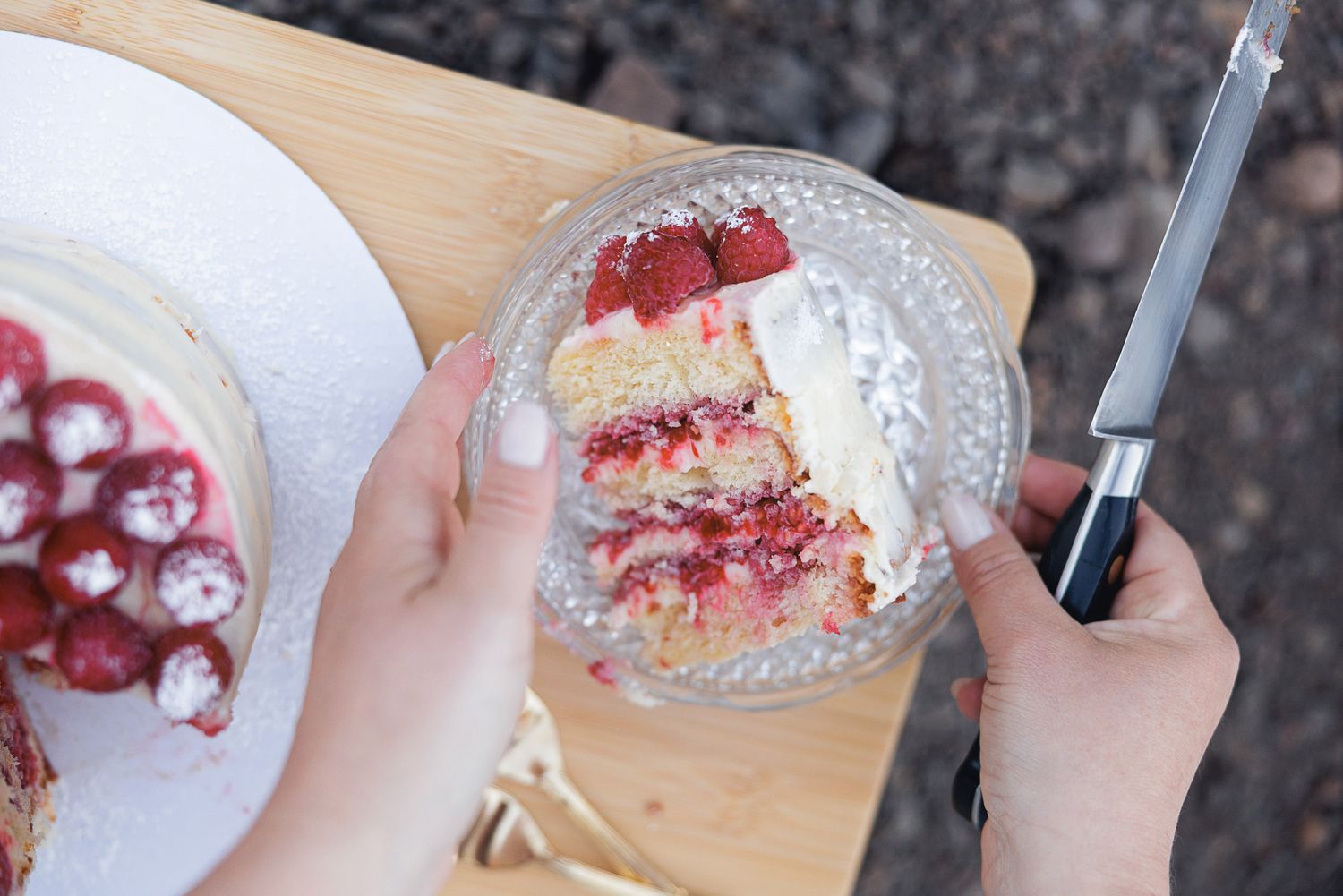Hands hold a glass plate with a slice of layered raspberry cake beside the whole cake, topped with fresh berries, on a wooden board.