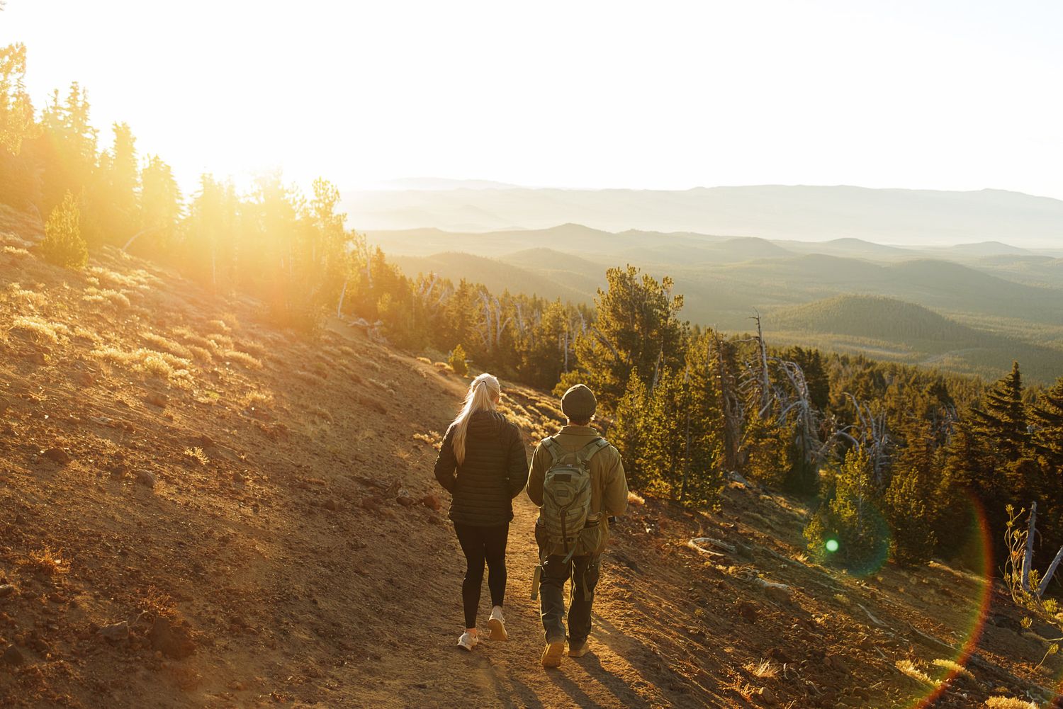 Two hikers walk down a sunlit mountain trail at golden hour, with evergreen trees and layered hazy ridgelines stretching into the distance.